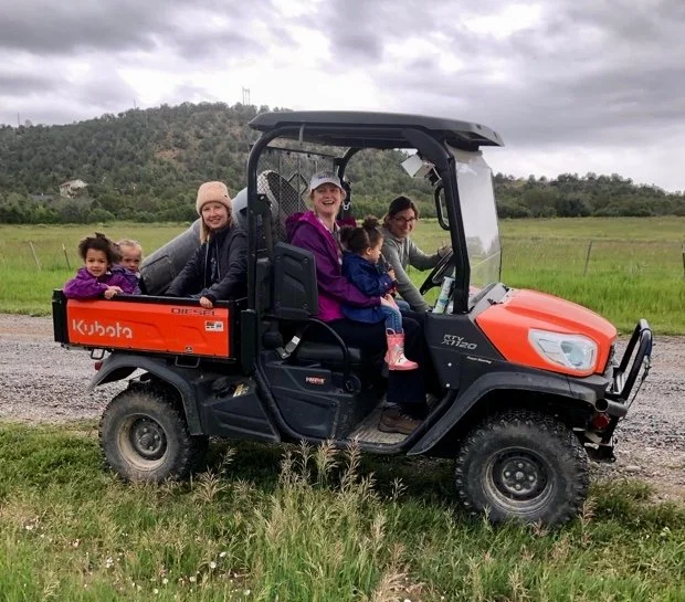 Multiple people, including children, riding in a Kubota utility vehicle on a grassy field with a cloudy sky and hilly landscape in the background.