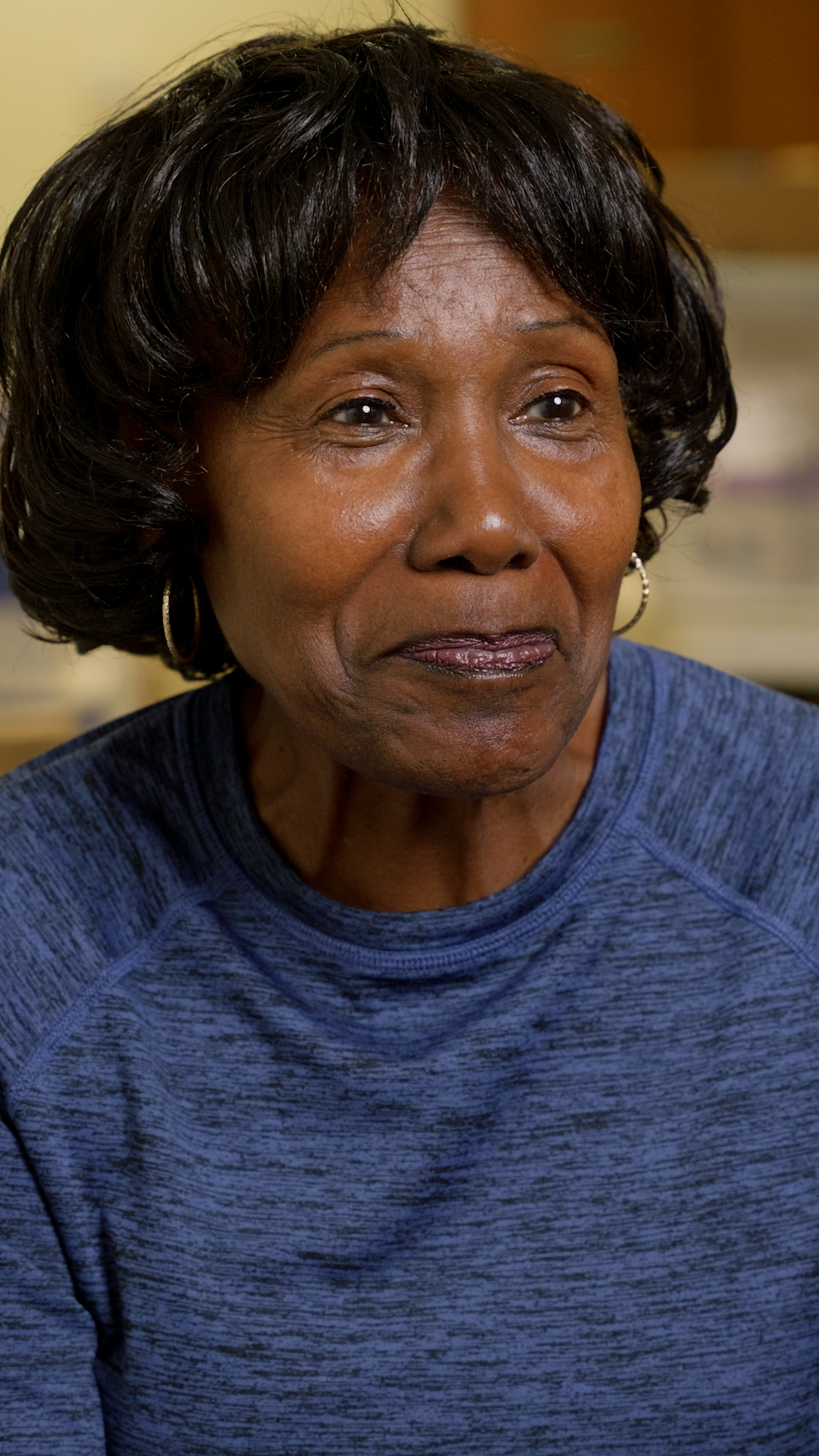 Close-up of an older woman with short, curly black hair, wearing a blue textured top and hoop earrings.