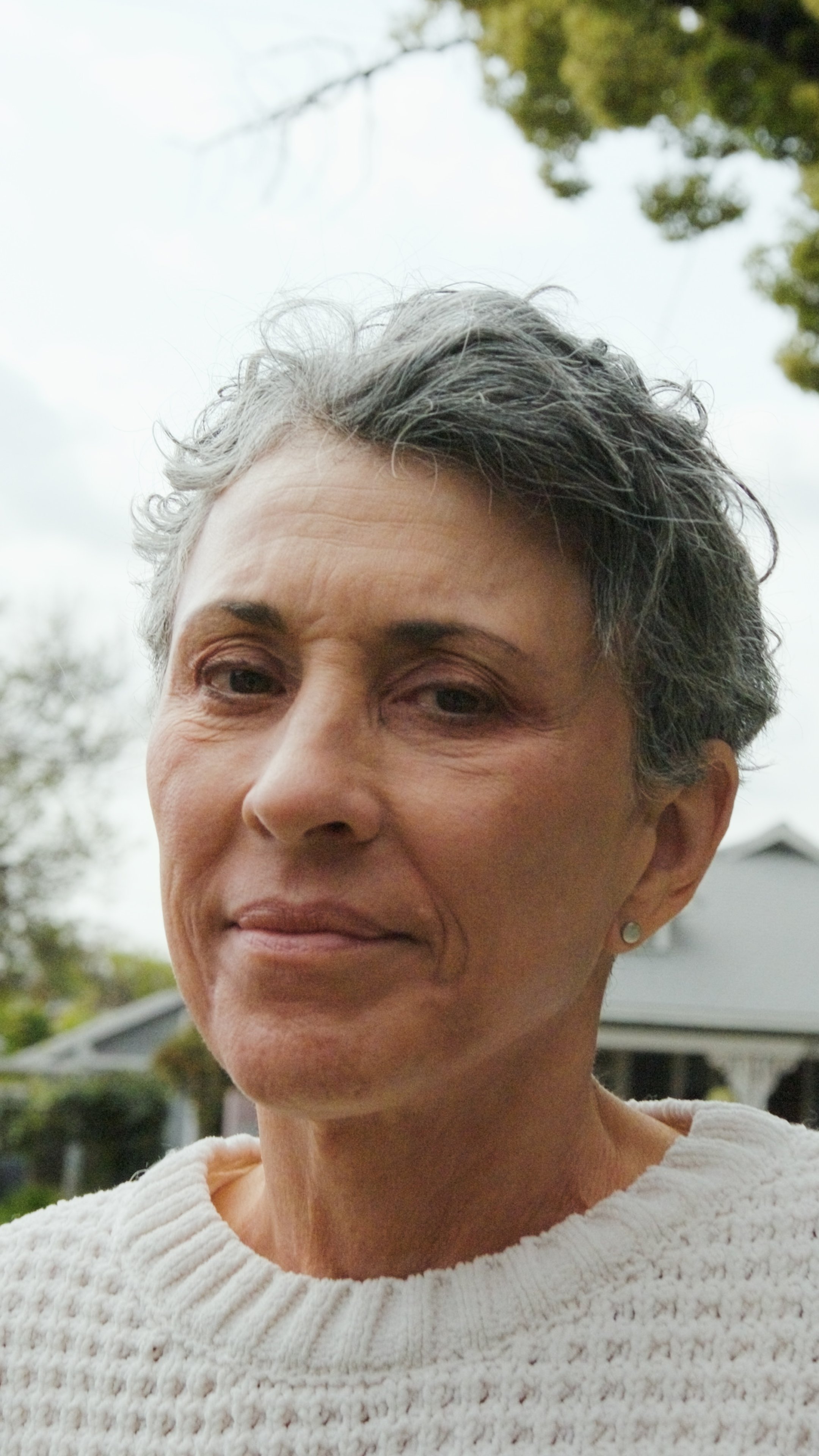Close-up of a middle-aged woman with short, curly gray hair wearing a white knit sweater, standing outdoors with trees and a house in the background.
