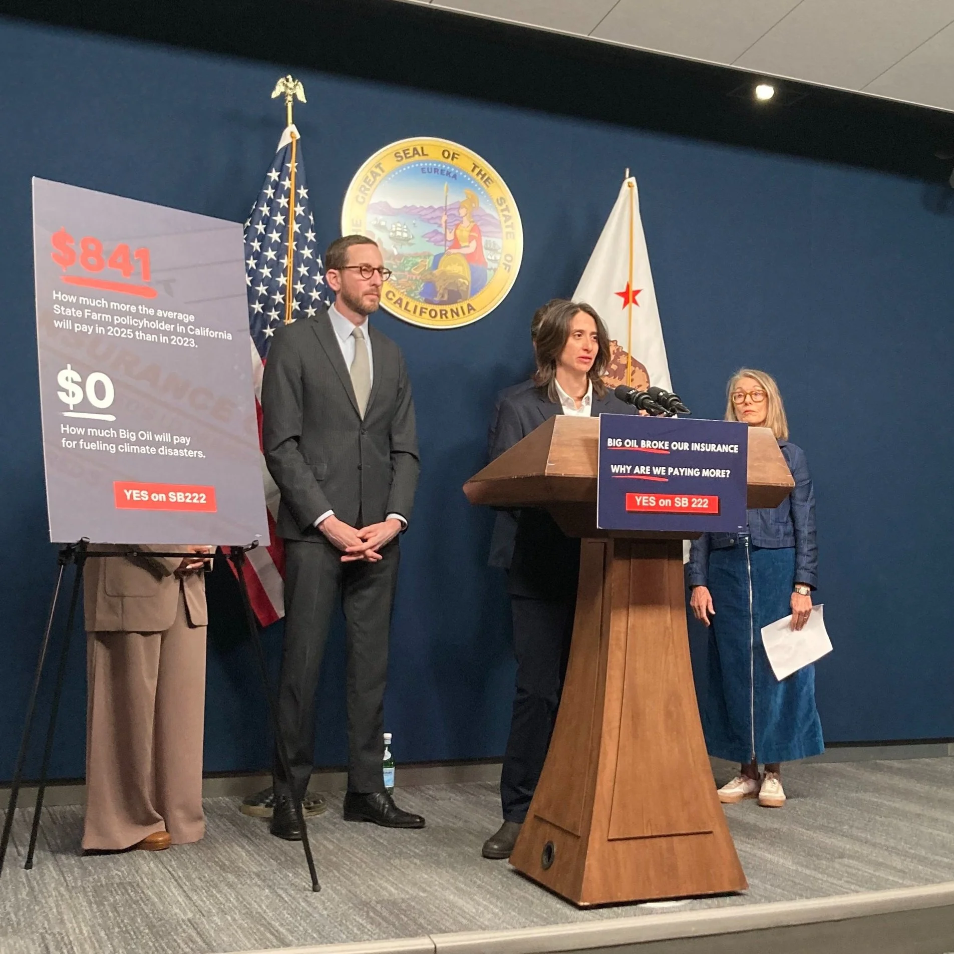 A woman is speaking at a podium during a press conference, with three other people standing behind her. There are signs discussing California's SB 222, with messages about insurance and oil companies.