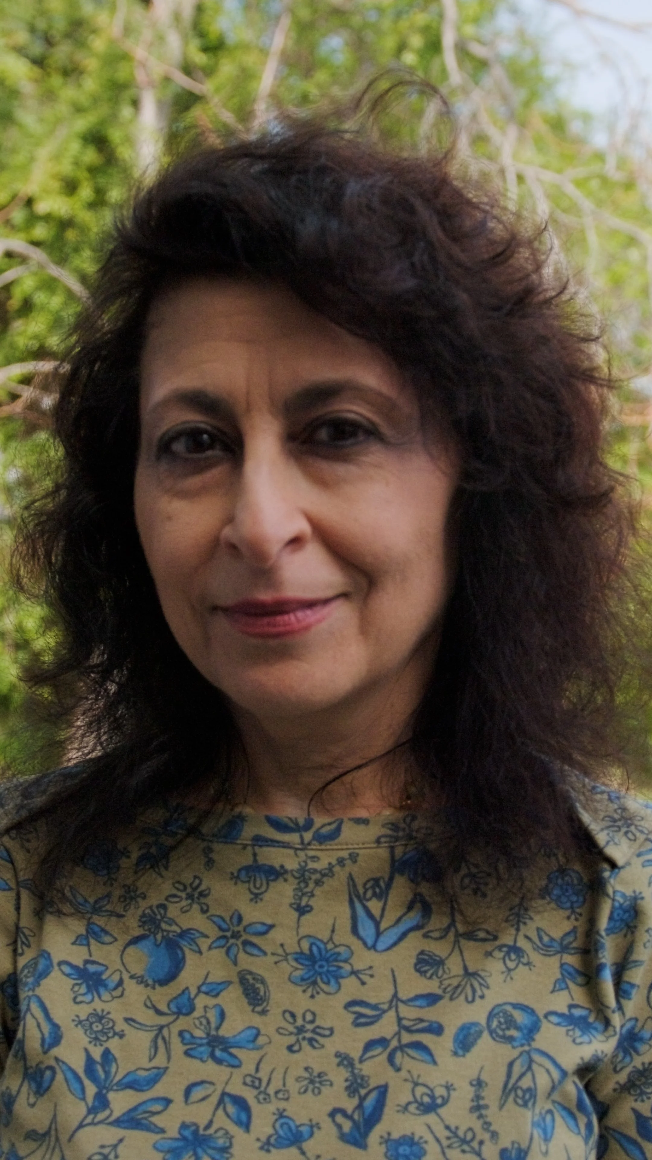 A woman with dark, curly hair standing outdoors with trees in the background, wearing a patterned shirt with blue floral and butterfly designs.
