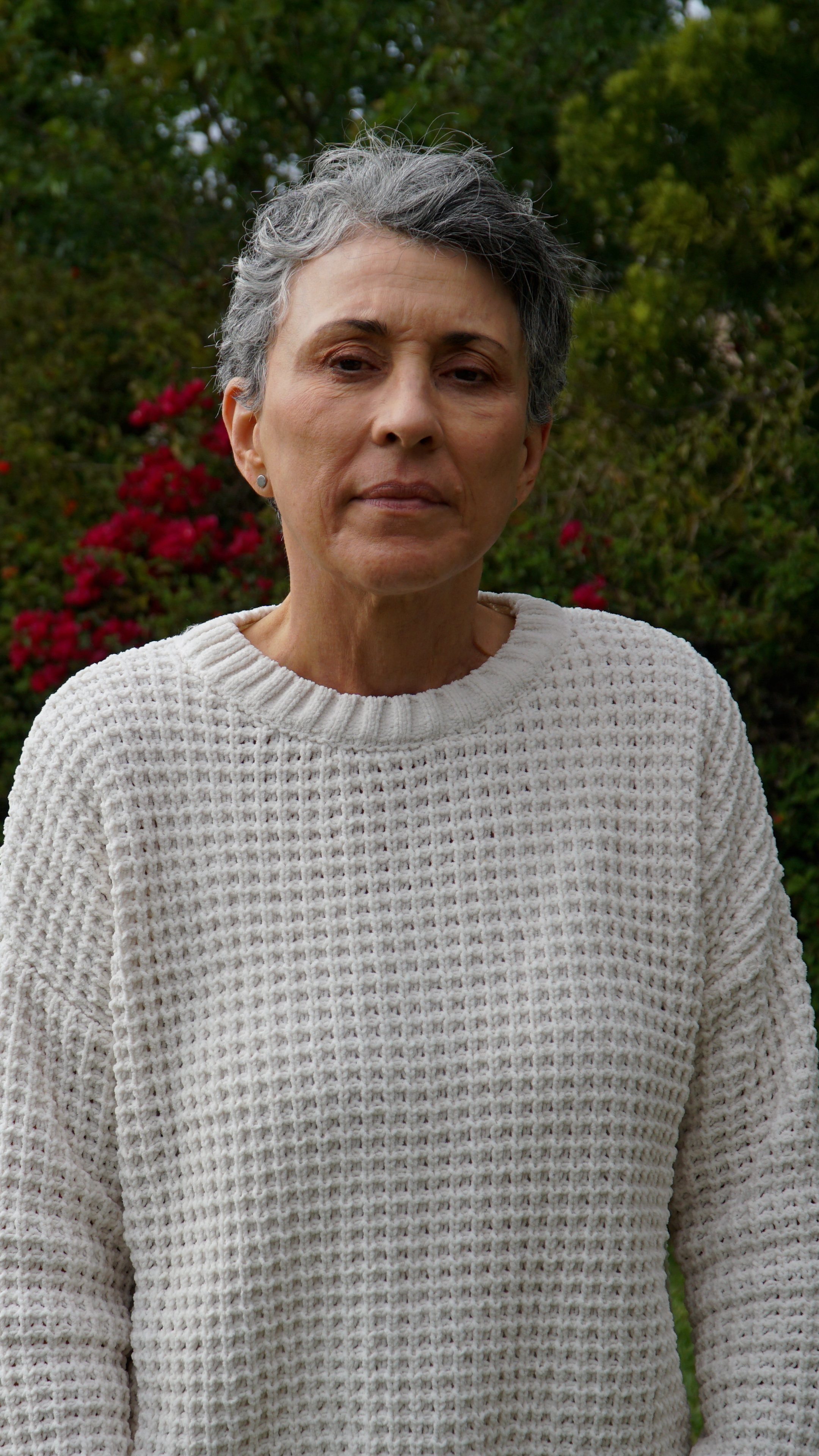 An older woman with short gray hair wearing a cream-colored knitted sweater standing outdoors with green trees and red flowers in the background.