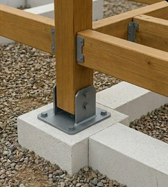 Esquina de una estructura de madera con soporte metálico anclado a bloques de cemento en un terreno con grava.