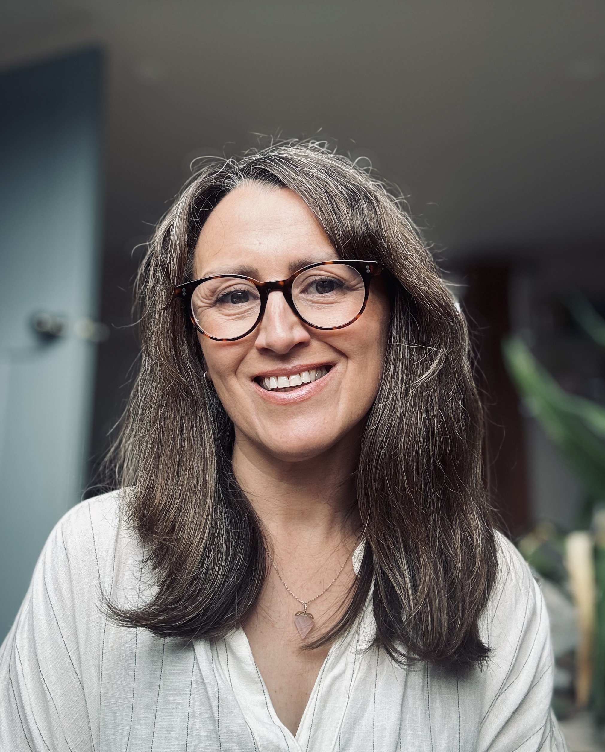 Close-up of a smiling woman with long dark hair, wearing glasses and a necklace, in an indoor setting.