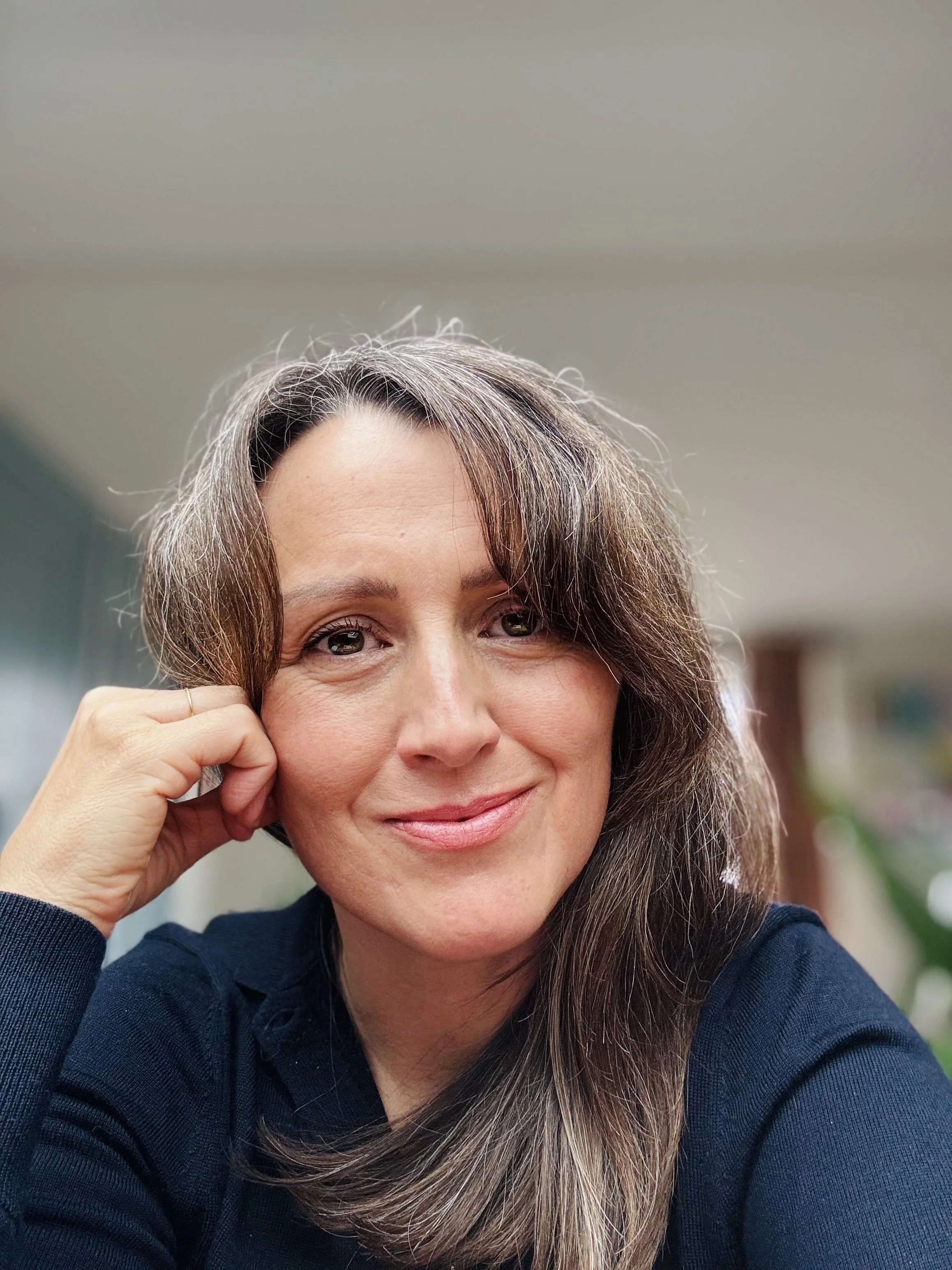 A close-up of a smiling woman with wavy brown hair, resting her chin on her hand, in a room with neutral walls.