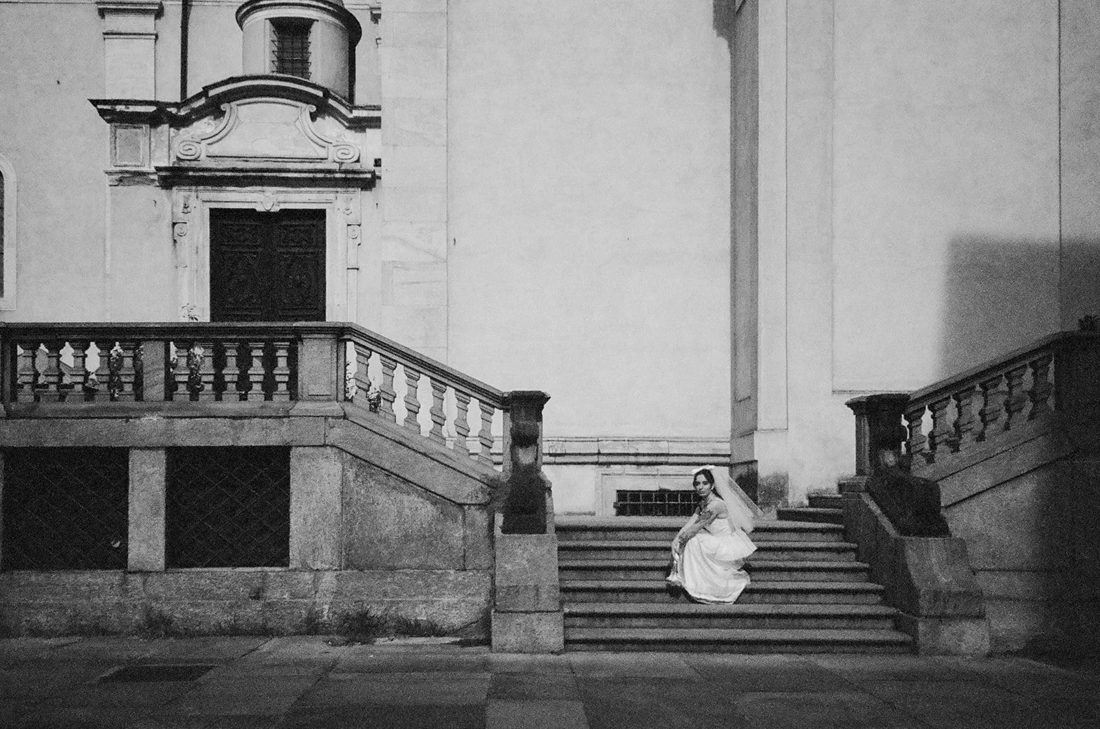 A woman in a long, white dress sitting on stairs outside a historic building with ornate architectural details in black and white.