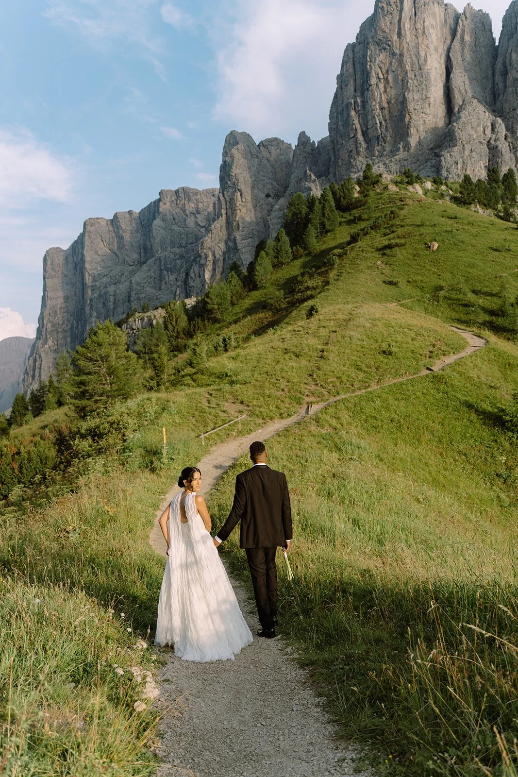 A bride and groom walking hand in hand on a narrow dirt path in a green mountainous landscape with tall rocky cliffs in the background.