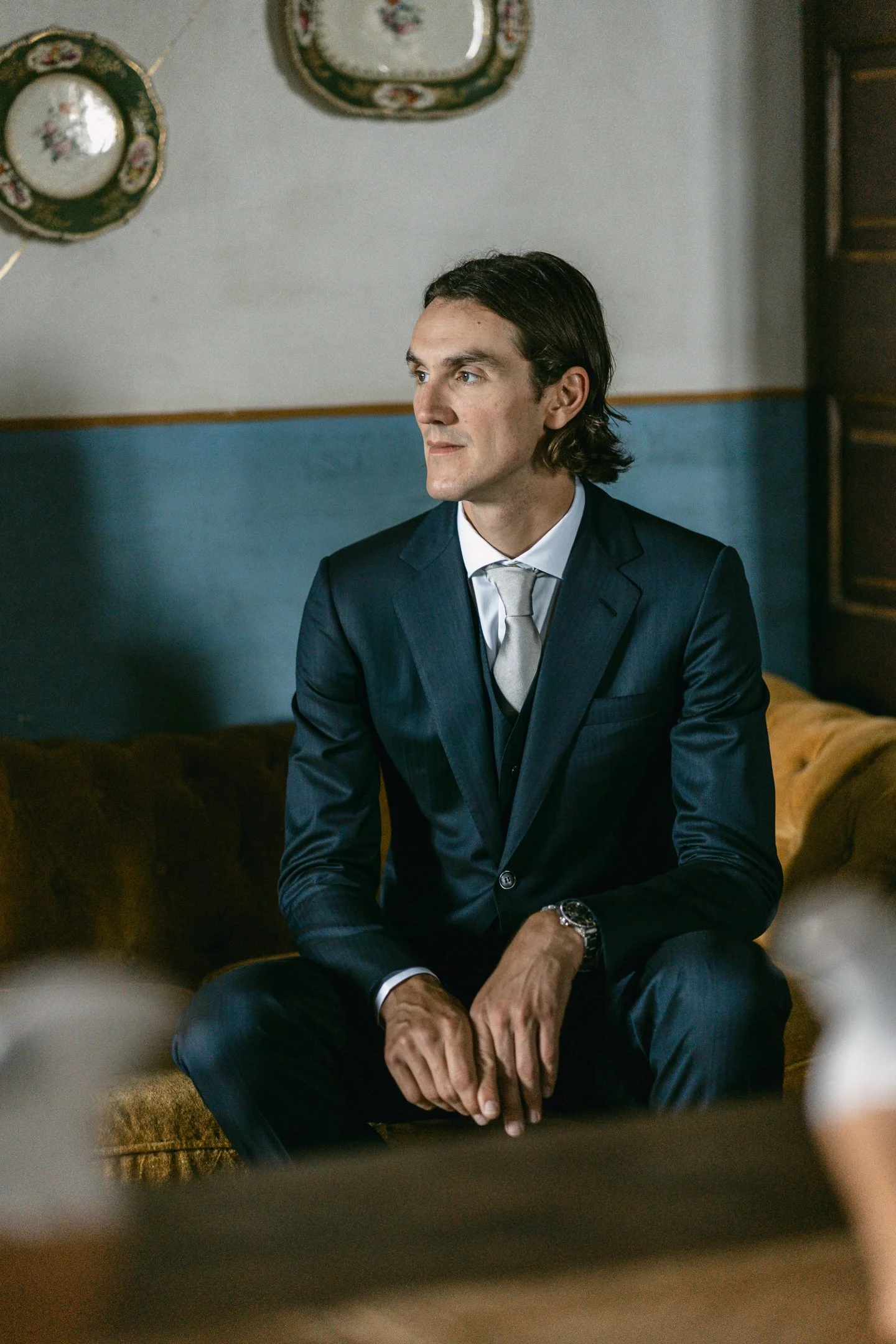 A man in a dark blue suit and silver tie sitting on a mustard-colored couch in a room with blue and white walls and decorative plates hanging on the wall.