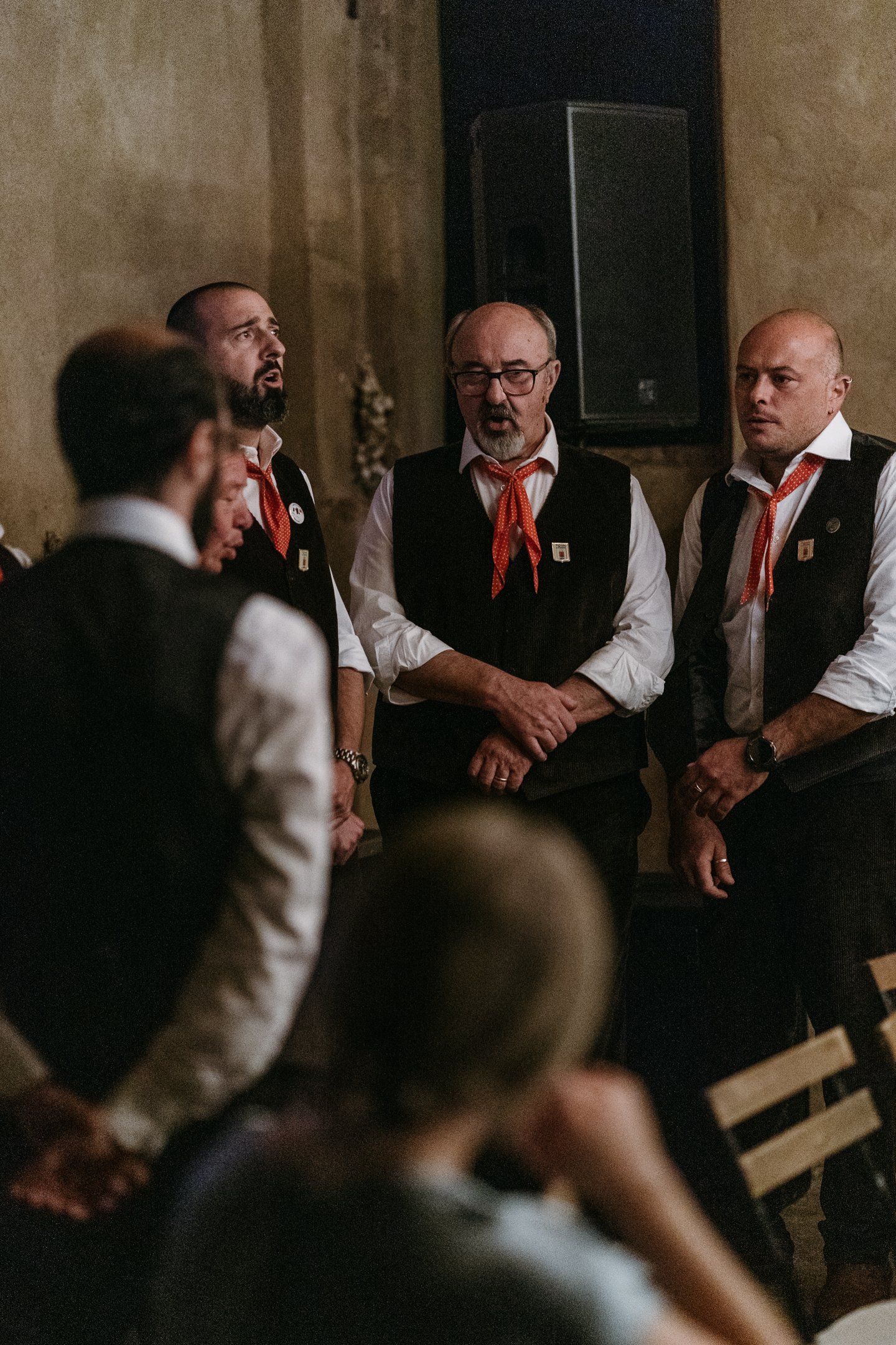 Group of men wearing black vests, white shirts, and red neckerchiefs, standing and singing together in a dimly lit room.
