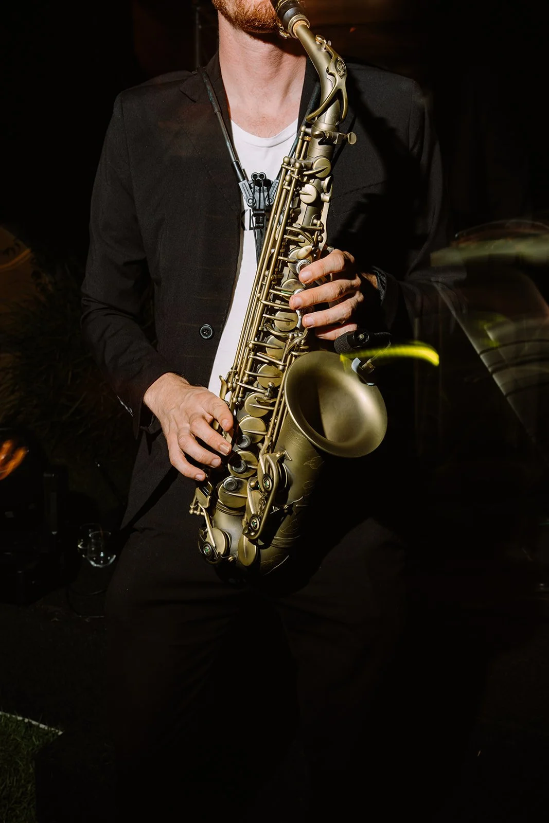 A musician wearing a black jacket and white shirt playing a gold-colored saxophone, with focus on the instrument and hands.
