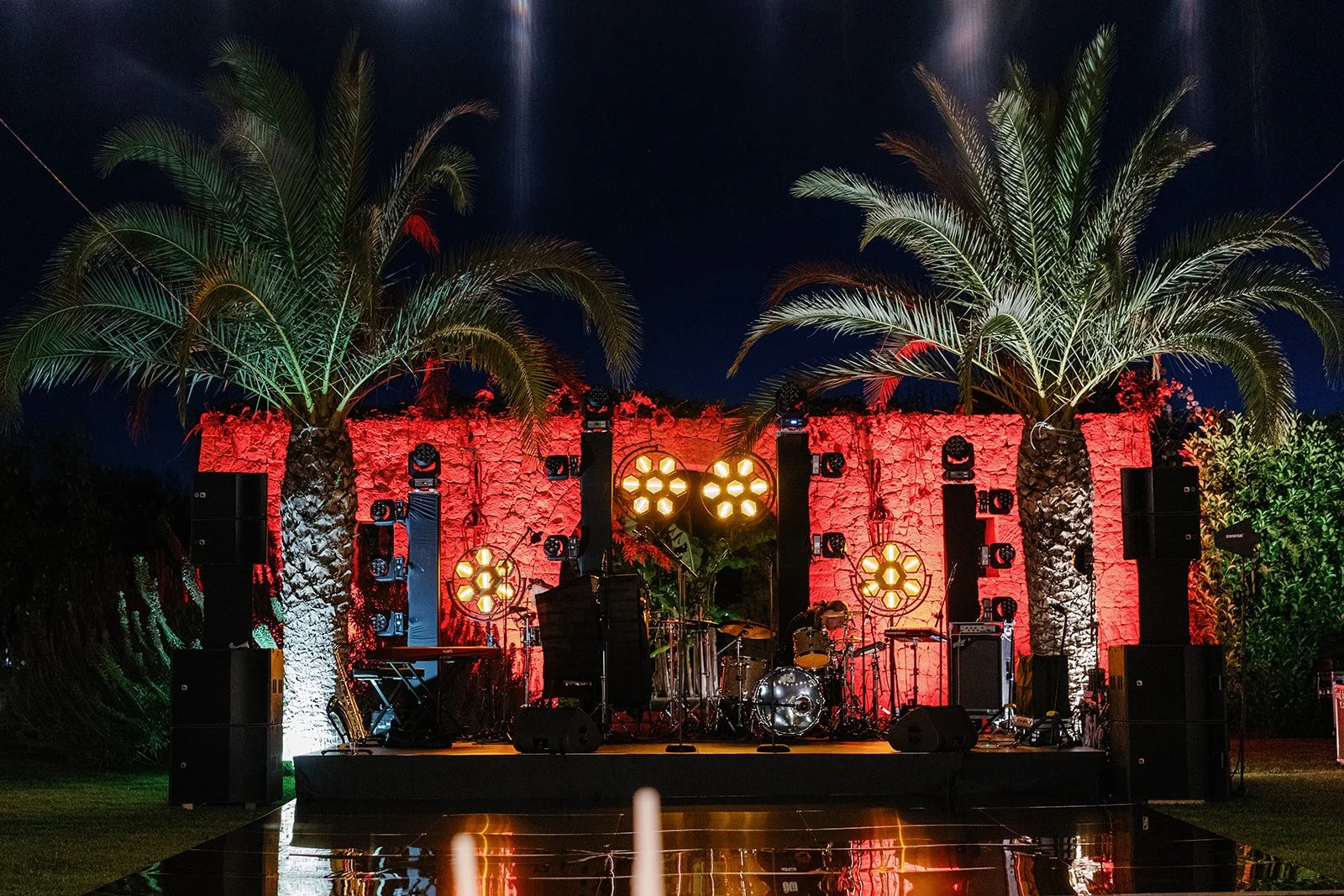 An outdoor stage set up for a performance at night, illuminated with yellow and red lights, with two large palm trees flanking the stage, and a red illuminated stone wall background.
