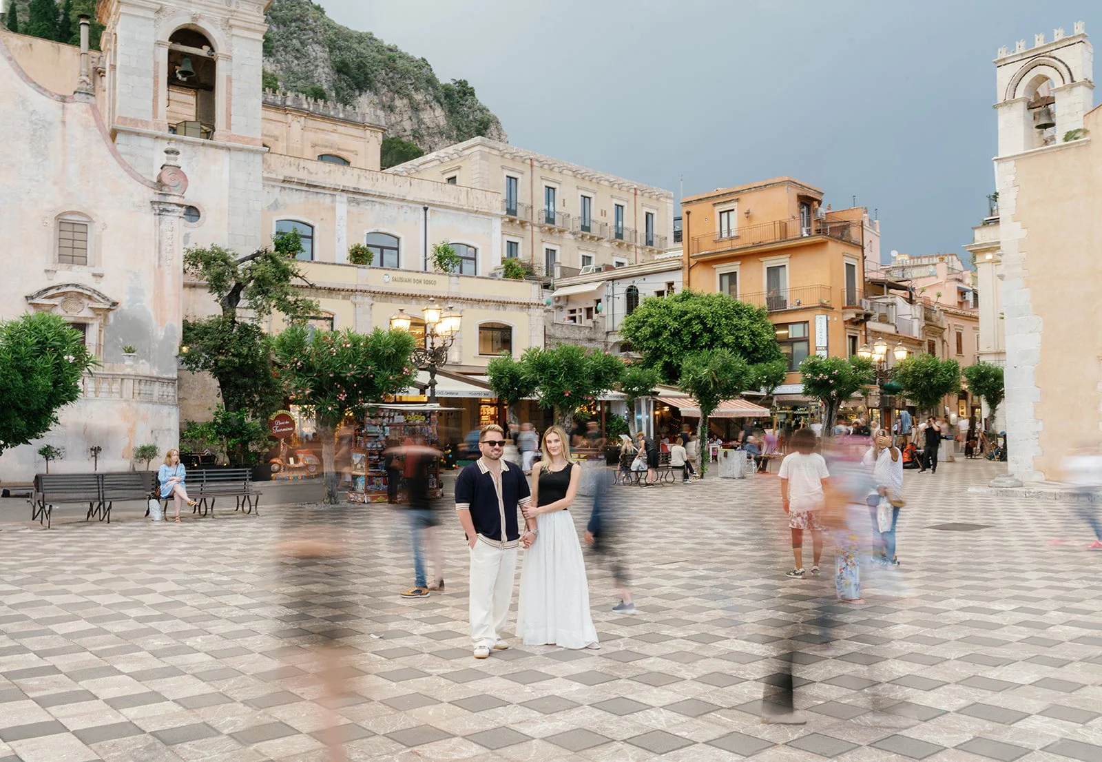 A couple holding hands standing in a lively town square with blurred pedestrians around them, colorful buildings, trees, and outdoor cafes in the background.