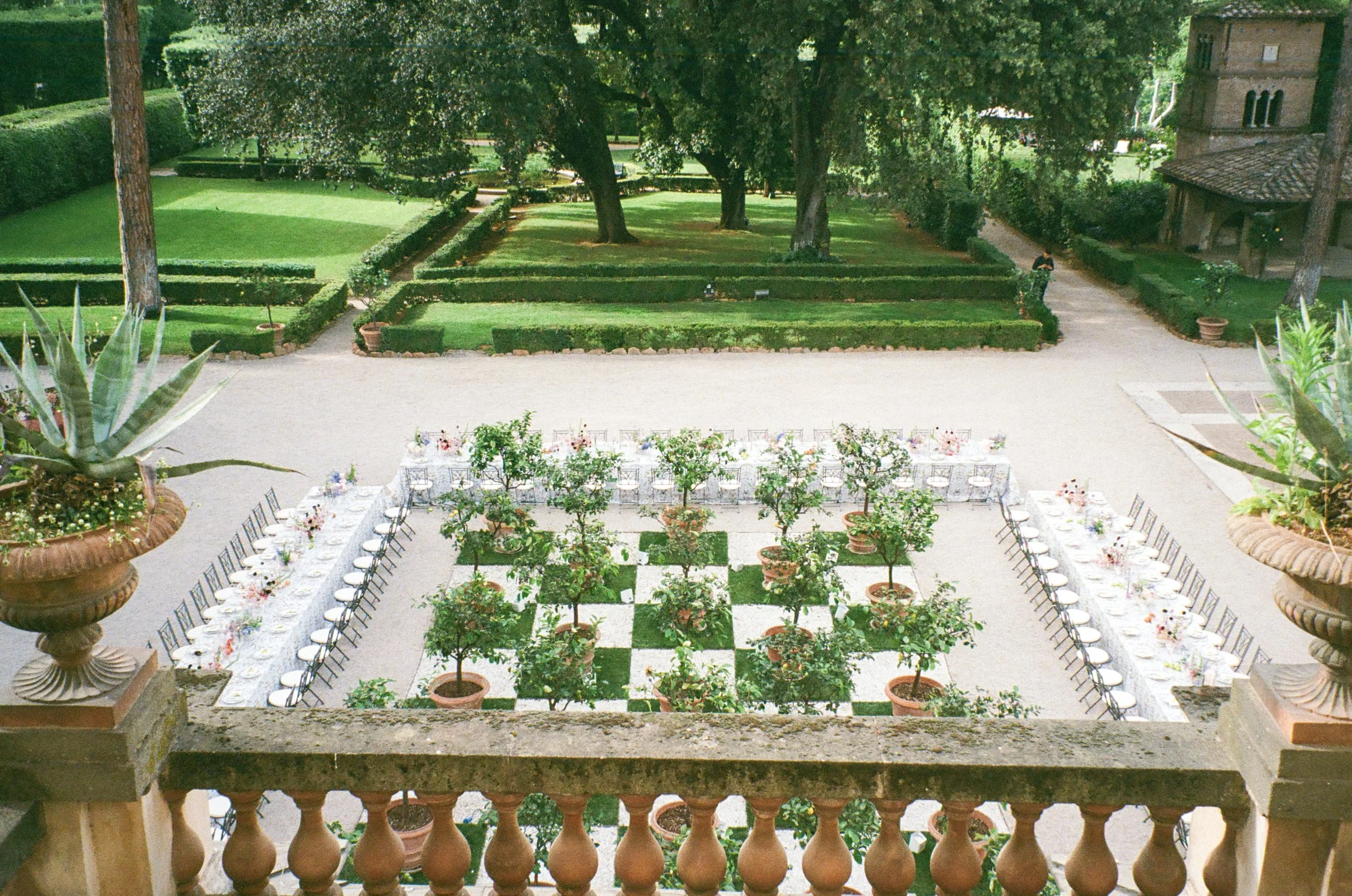 A formal outdoor dining setup arranged on a checkered lawn with potted plants, surrounded by lush green hedges and trees in a garden or estate.