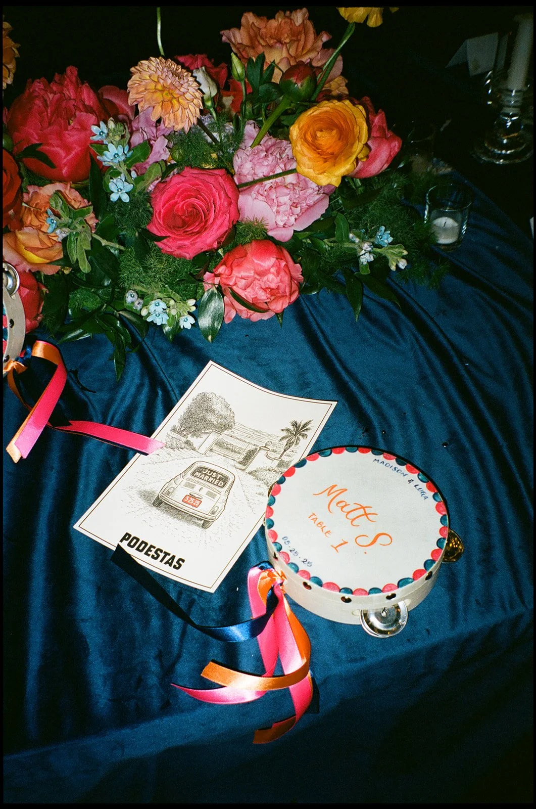 A floral centerpiece with pink, red, yellow, and peach flowers on a dark blue tablecloth. A white round sign with red, white, and blue decoration, reading 'Matt S., Table 1,' is on the table. A black and white drawing of a car with a 'Just Married' s