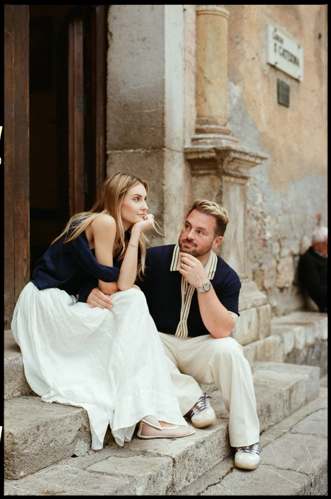 A young woman and man sitting on stone steps outside a rustic building, engaged in a casual conversation, with the woman resting her chin on her hand.