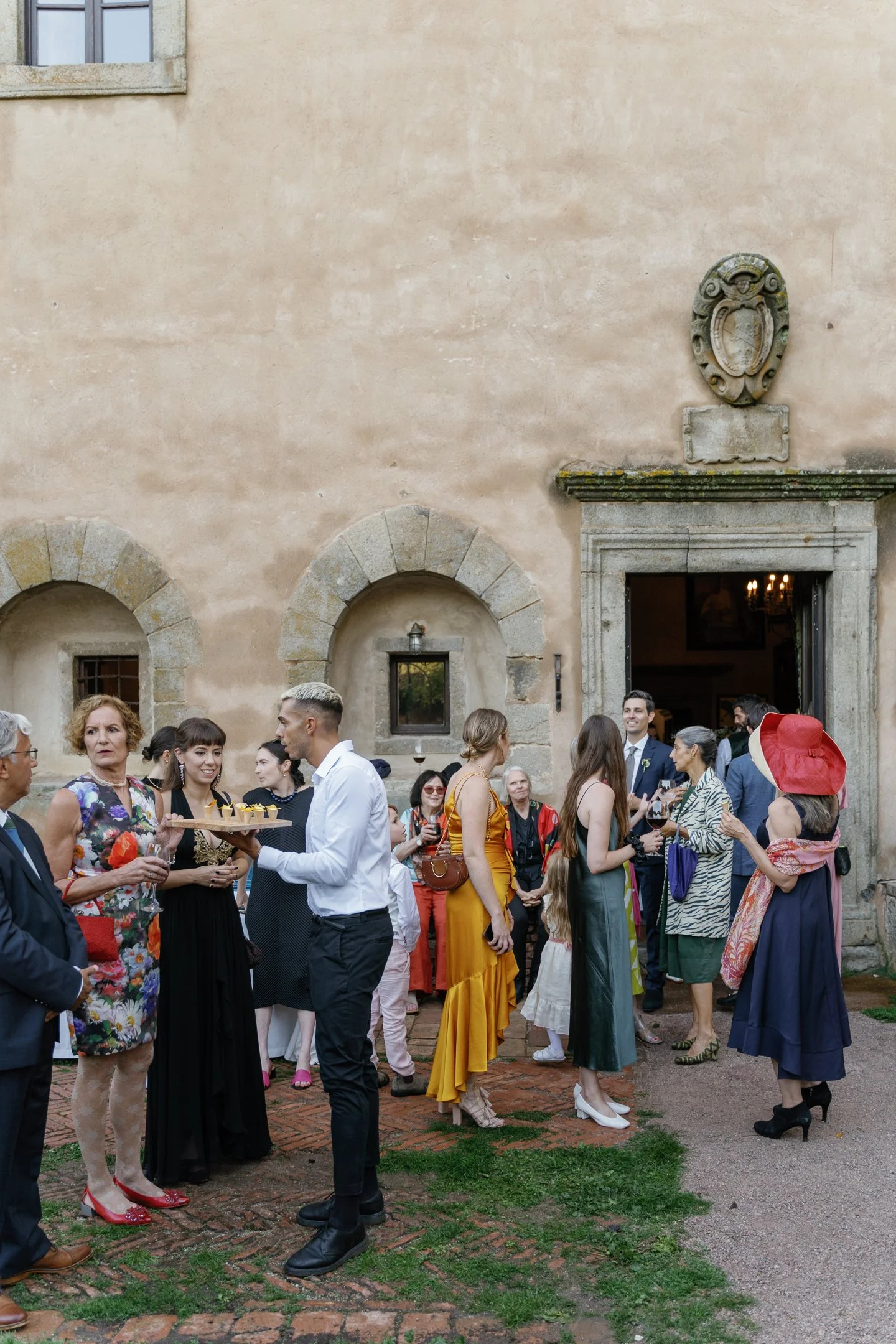 People gathered outside a rustic building celebrating a festive event, with some holding drinks and others socializing, against a historic stone wall.