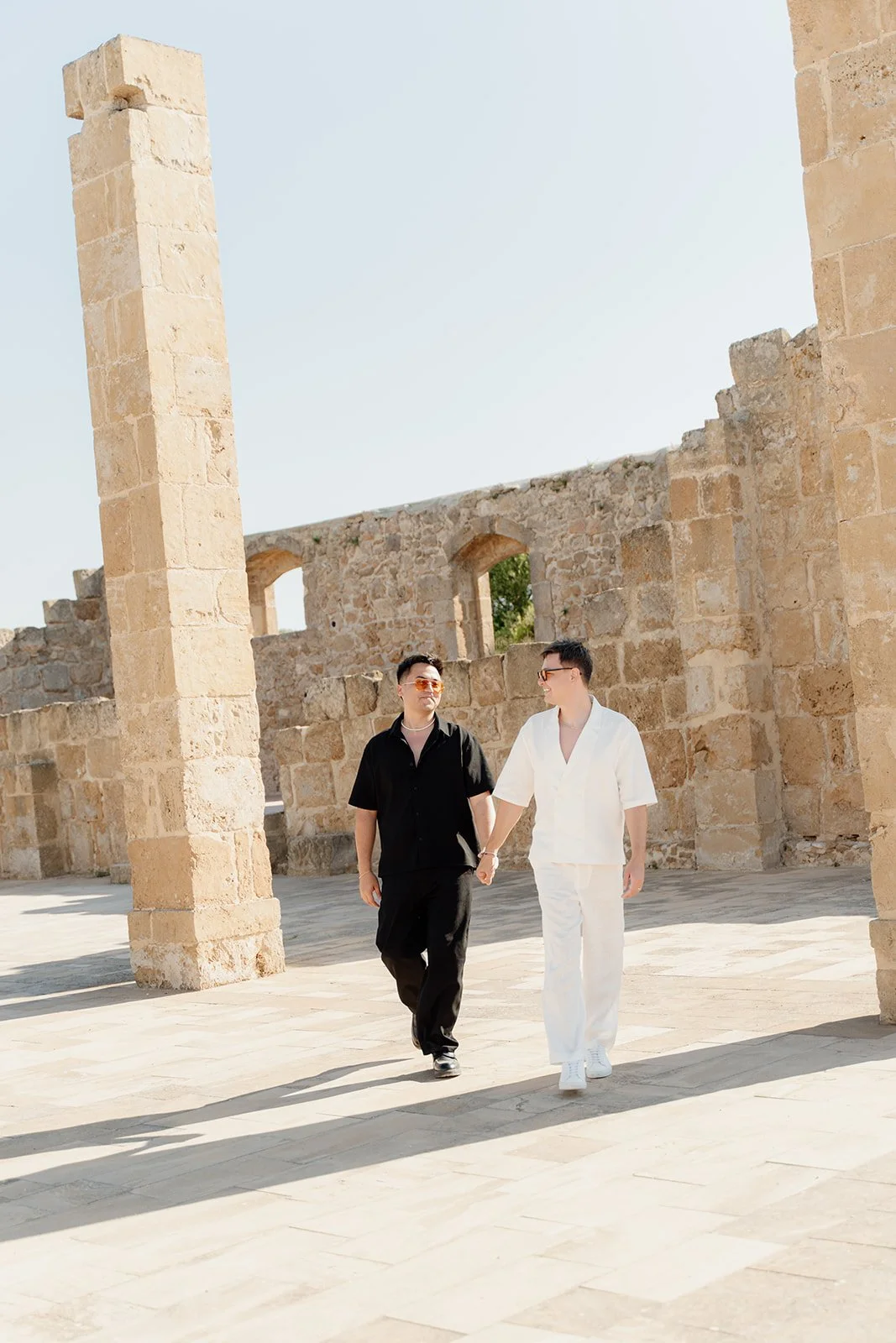 Two people holding hands and walking through an ancient stone ruins under a clear sky, one in black and the other in white clothing.