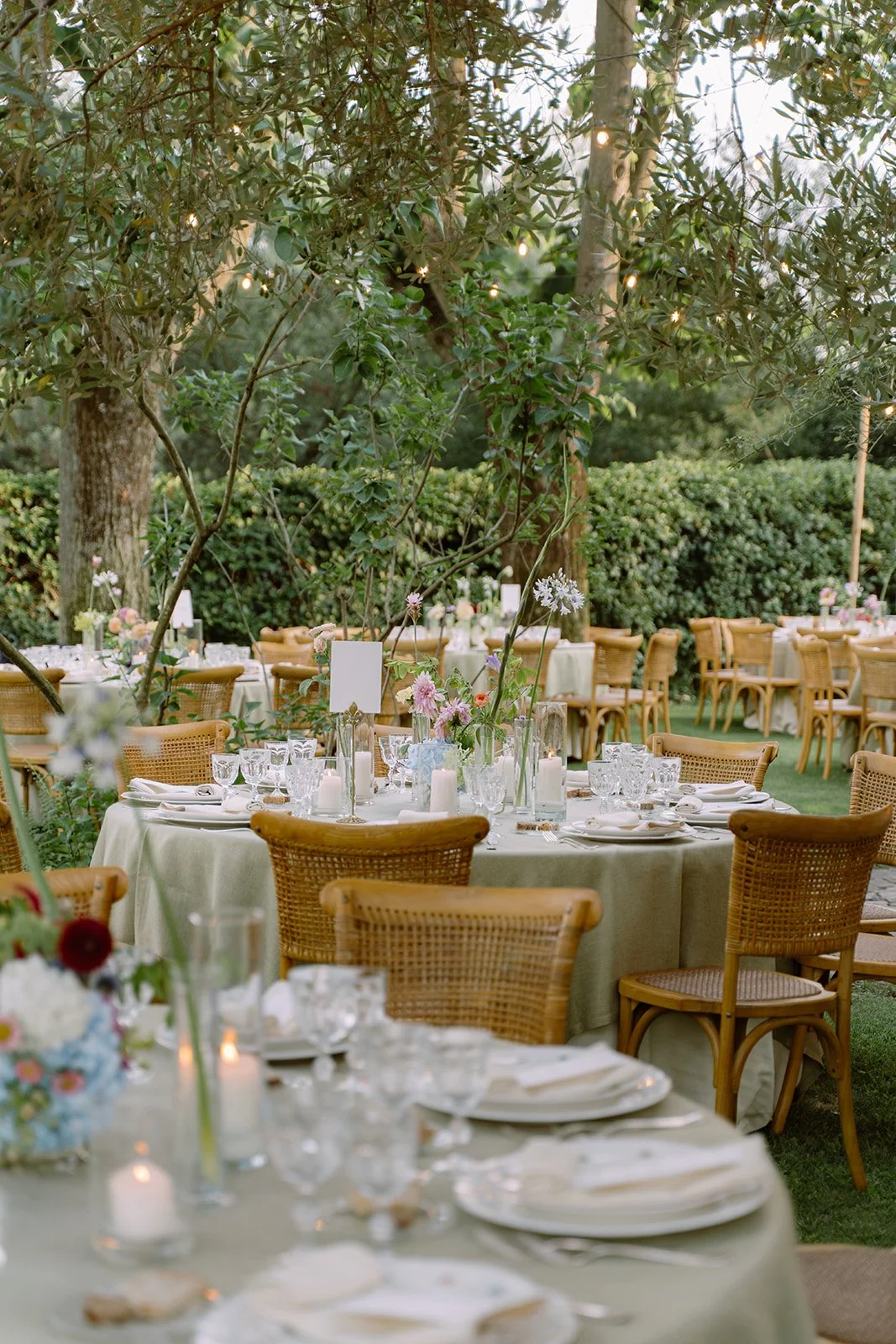 Outdoor reception area with round tables covered in light-colored tablecloths, decorated with flowers, candles, and glassware, set beneath trees with string lights.