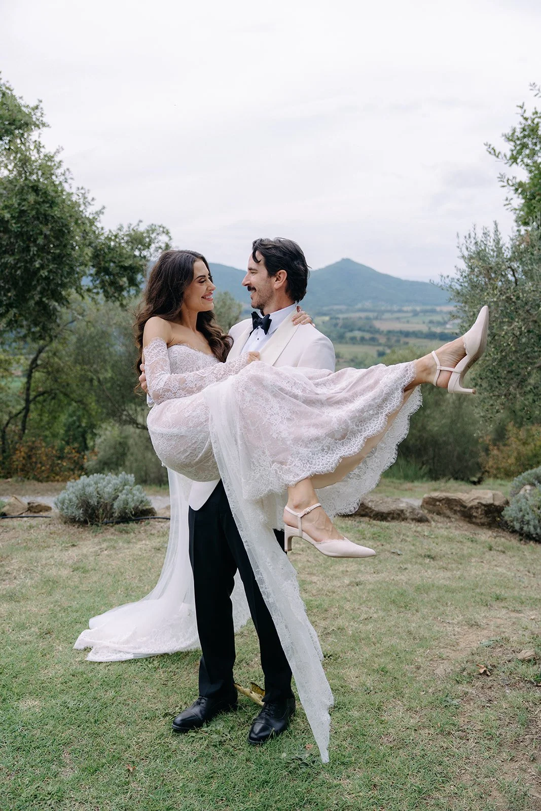 A groom in a tuxedo carrying a bride in a lace wedding dress outdoors, with trees and mountains in the background.