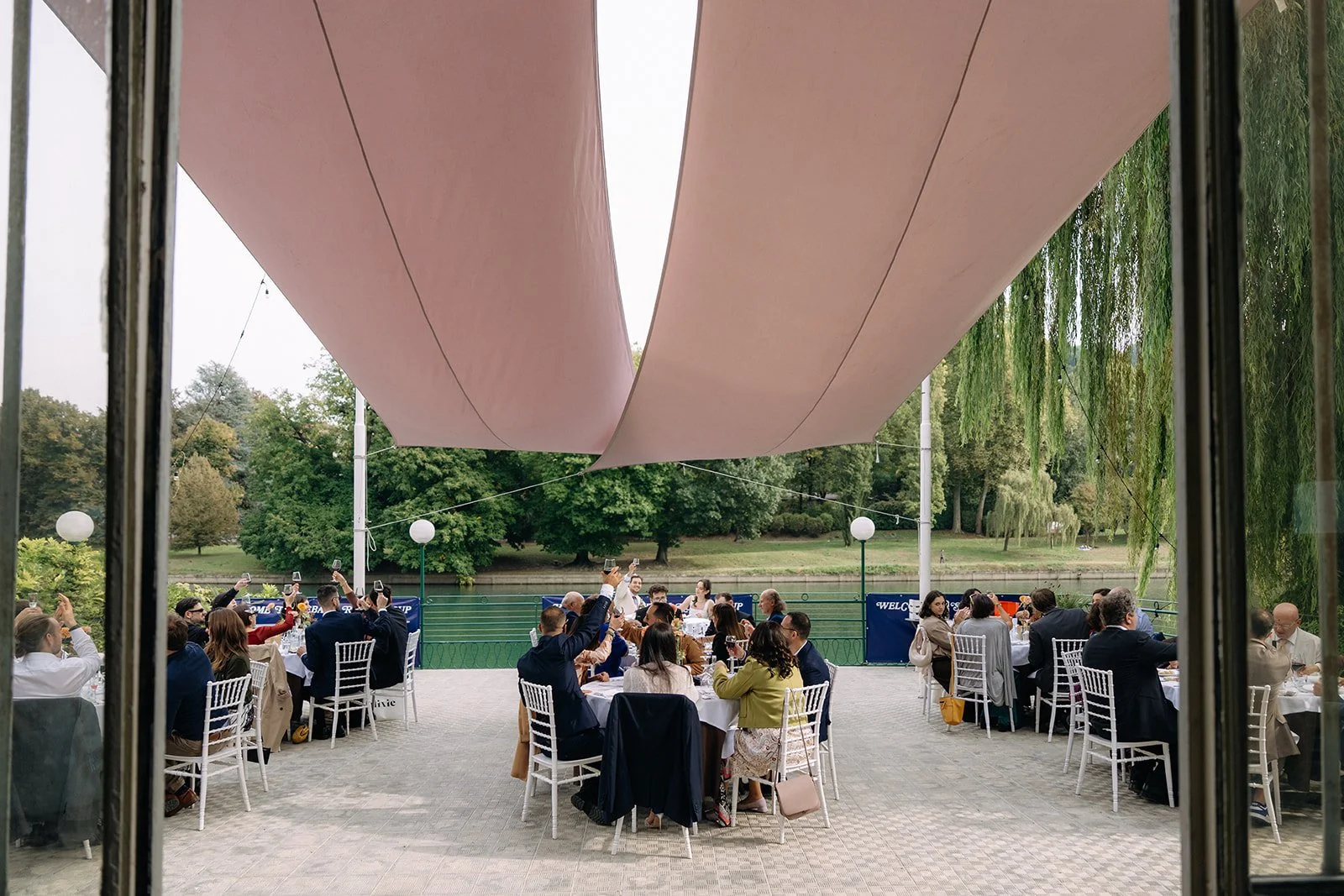 People dining at outdoor tables by a river, with large pink canopy overhead, trees in the background, and lantern-style lights.