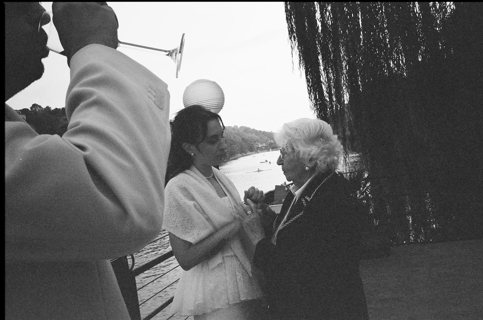 A black-and-white photo of a wedding scene by a river. The bride and an elderly woman stand close, holding hands and gazing at each other. Two men, one with a wine glass and the other holding a champagne flute, stand nearby. The background features t