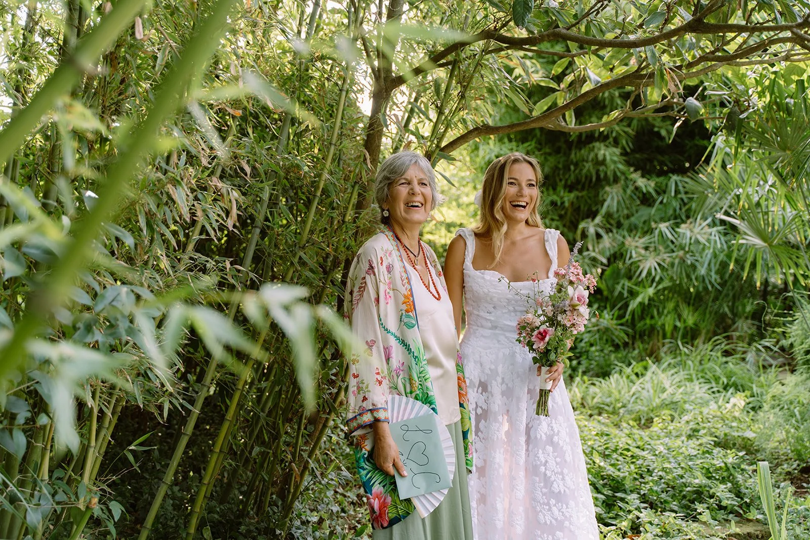 A bride in a white lace wedding dress holding a bouquet standing next to an older woman in a colorful floral outfit and holding a decorated fan, both smiling in a lush green outdoor setting.