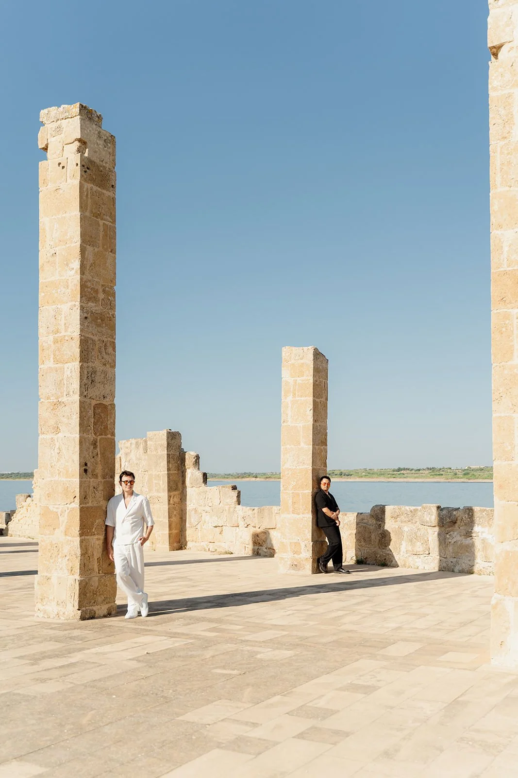 Two people, a woman in white and a man in black, stand near ancient stone ruins by a body of water under a clear blue sky.