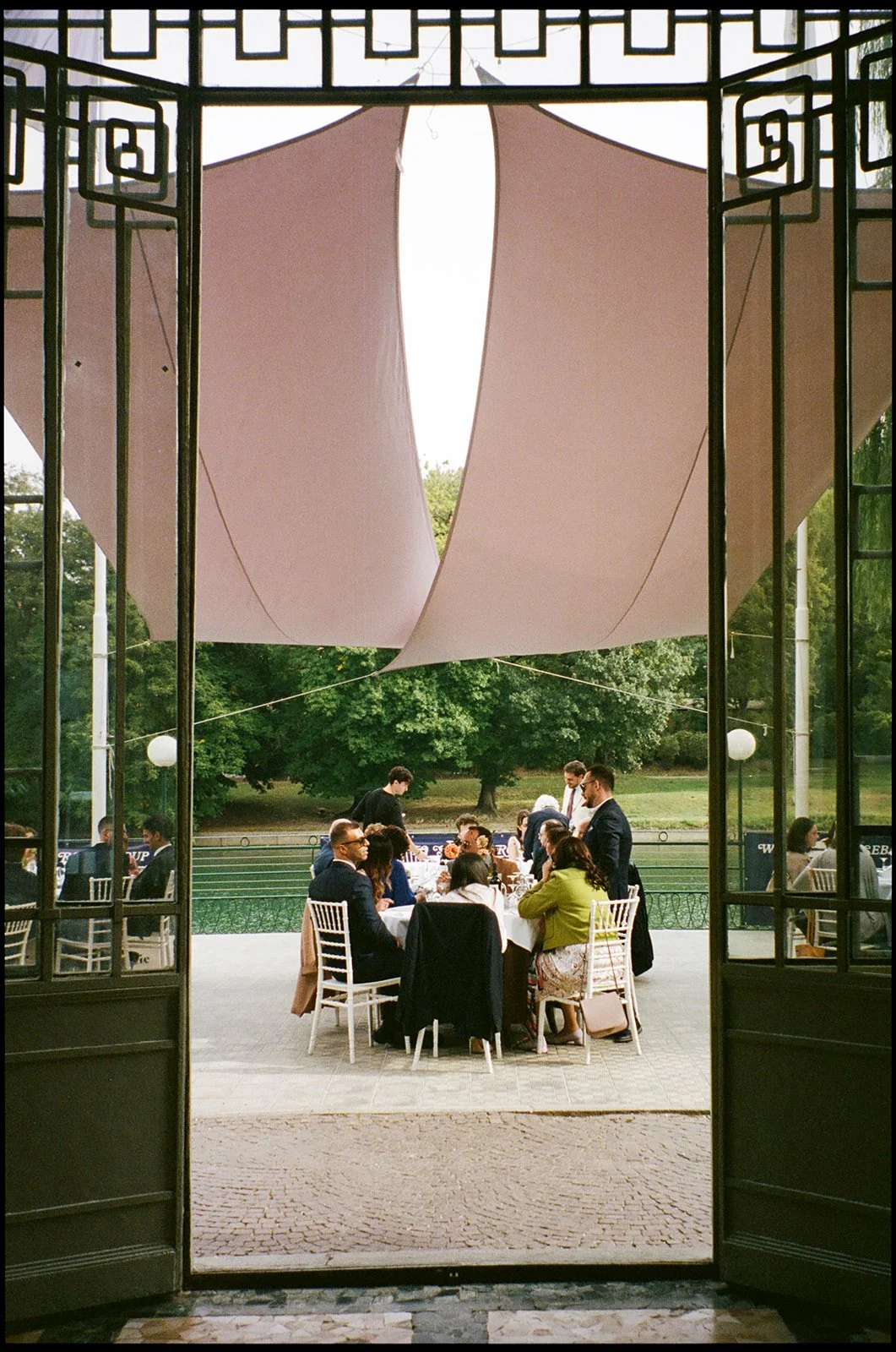 People dining outdoors at a formal event under a large fabric canopy, seen through open doors.