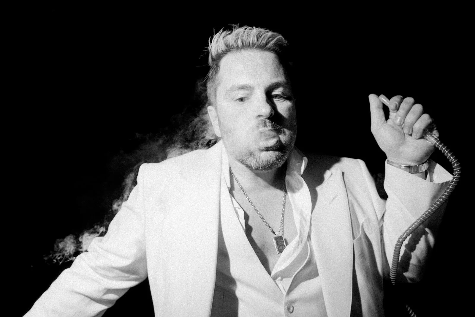 A black and white photo of a man with sunglasses, a beard, and styled hair, wearing a white suit and holding a vintage microphone.