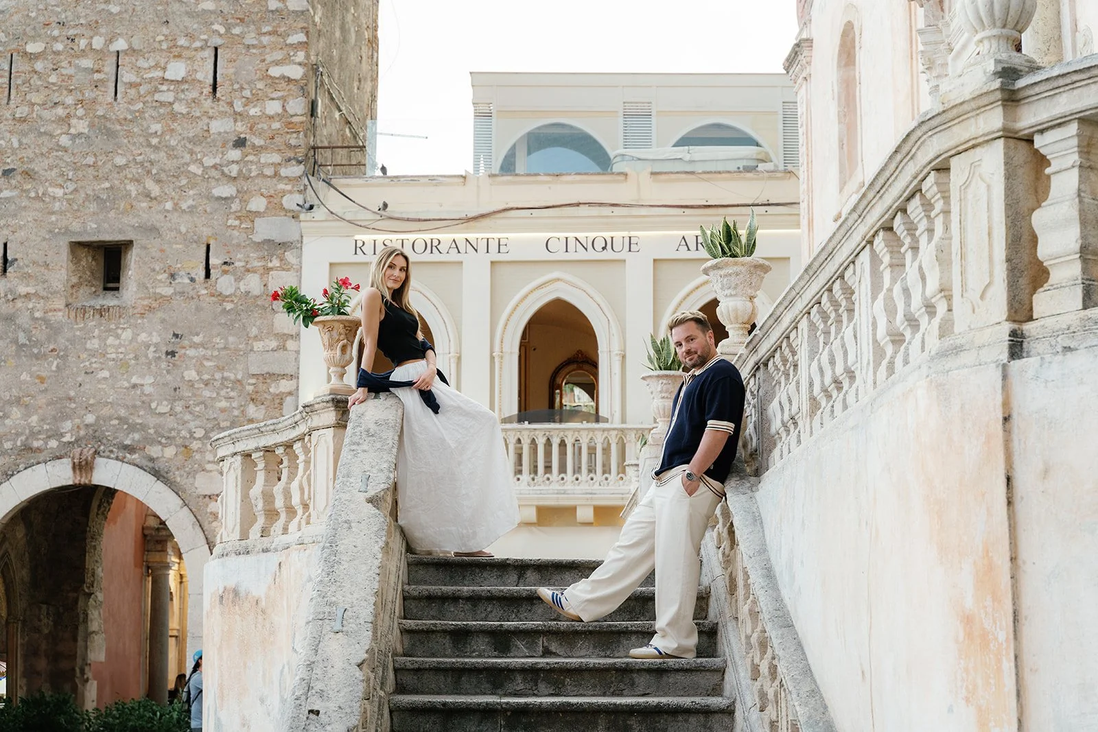 Two young adults, a woman and a man, pose on a stone staircase outside a European style building with the words 'Ristorante Cinque' above the entrance. The woman is sitting on the ledge, wearing a black tank top and a white skirt, with a black sweate