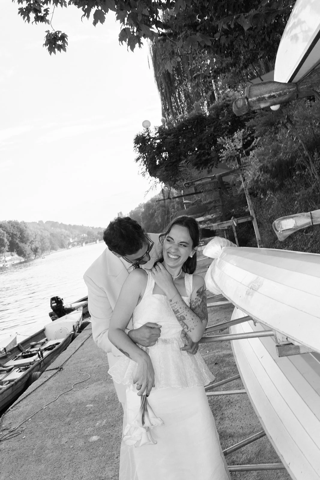 A couple dressed in wedding attire sharing a joyful moment by the river, with boats, trees, and a lakeside scene in the background.