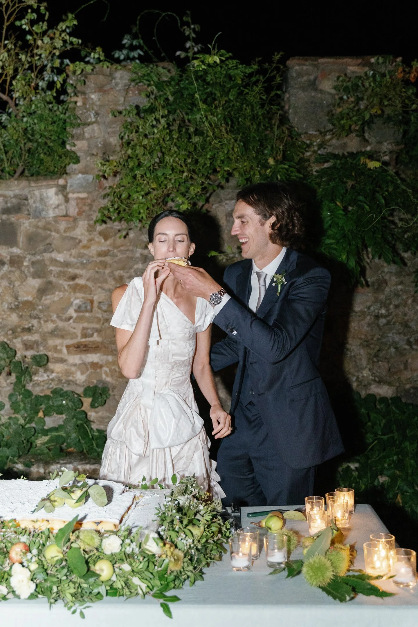 A newlywed couple sharing a slice of wedding cake outdoors at night.