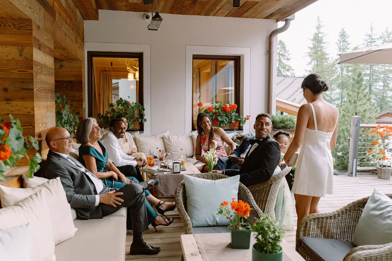 Group of adults and children enjoying a gathering on a patio with wooden decor, flowers, and a mountain view.