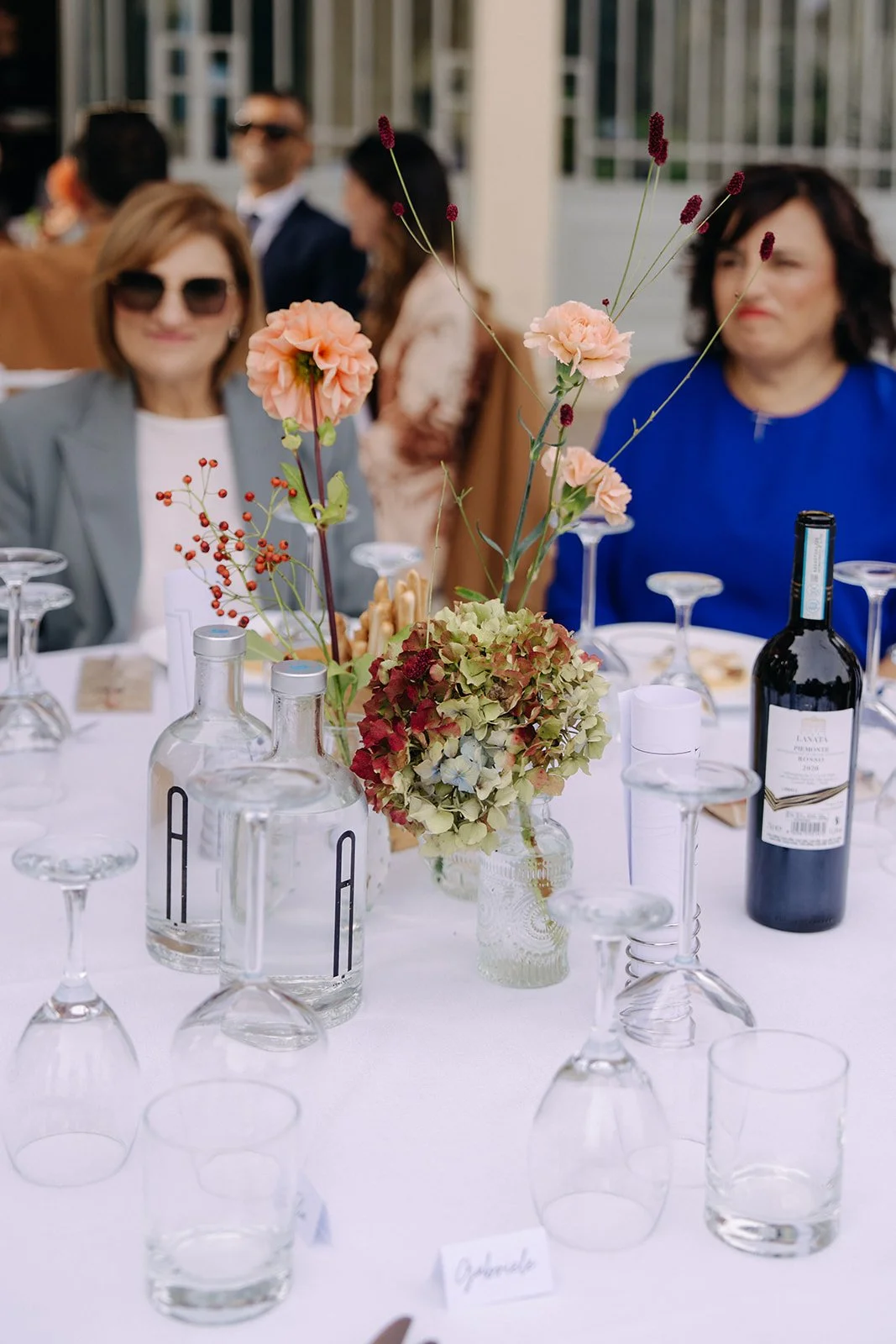 Table decorated with a floral centerpiece, wine bottle, and glassware at a social gathering with people in the background.