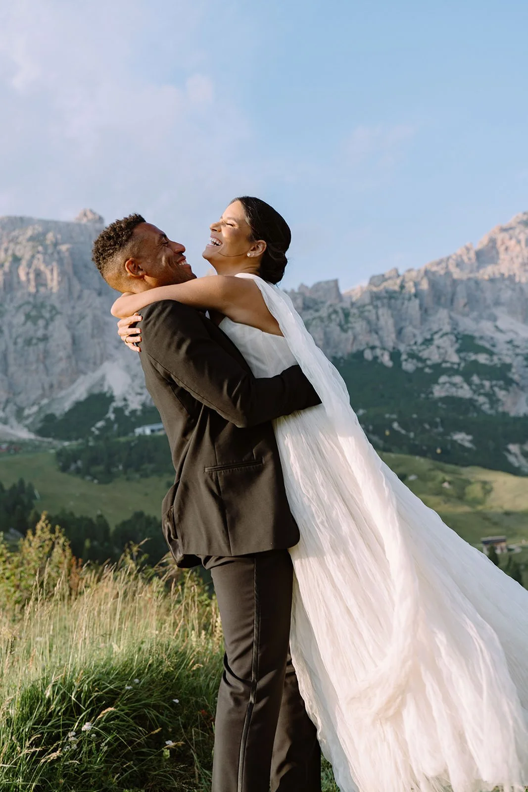 A happy couple in wedding attire, embracing outdoors with mountains and blue sky in the background.