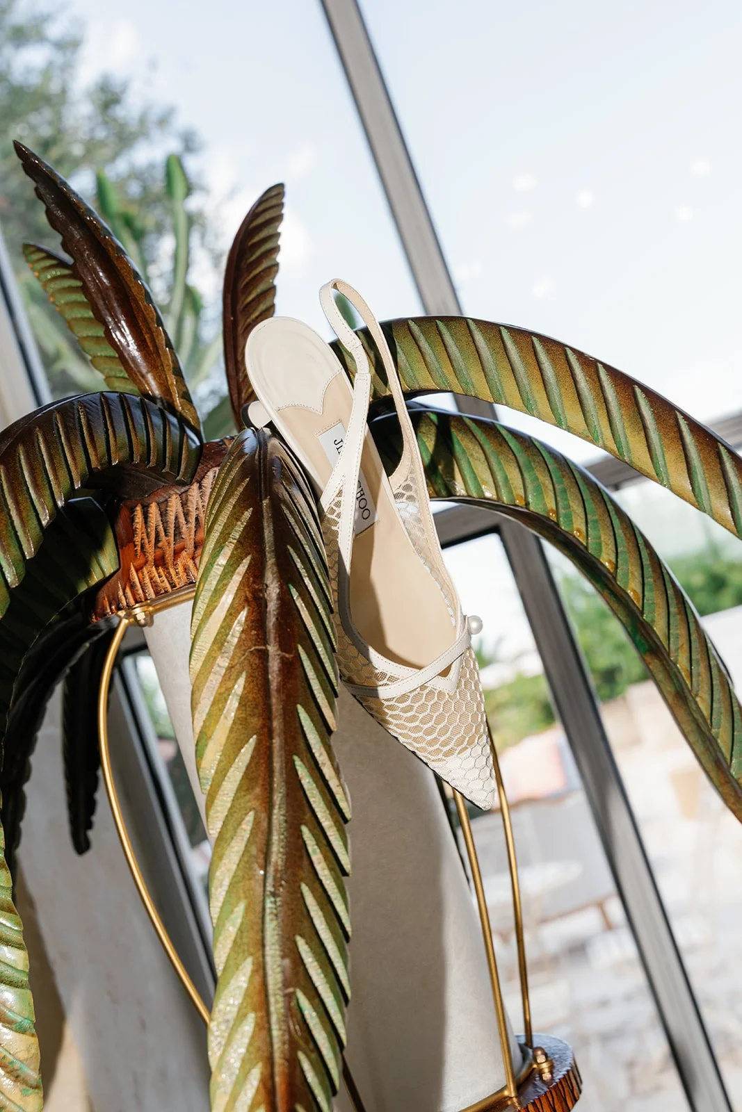 A decorative plant with large green and brown leaves hangs on a gold plant stand, with white high-heeled shoes with mesh design resting on the leaves. There is a window in the background with a view of trees and a cloudy sky.