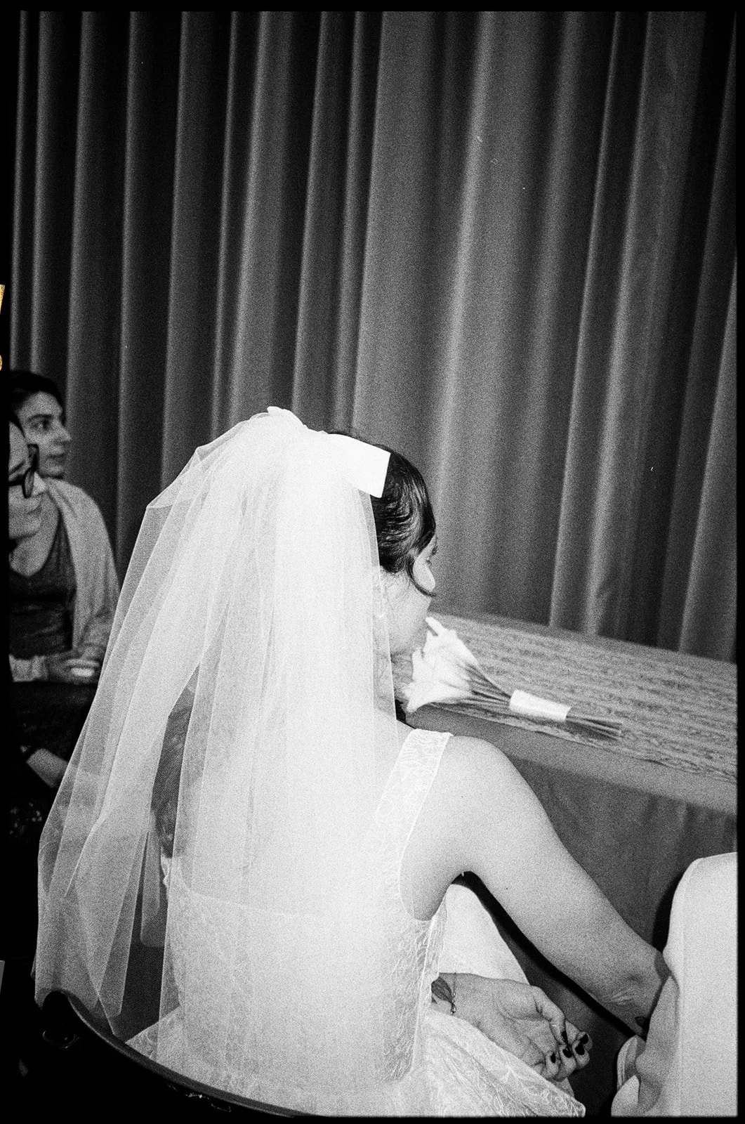 A bride wearing a veil sitting at a table with a bouquet of flowers, with a woman visible in the background at what appears to be a wedding reception.