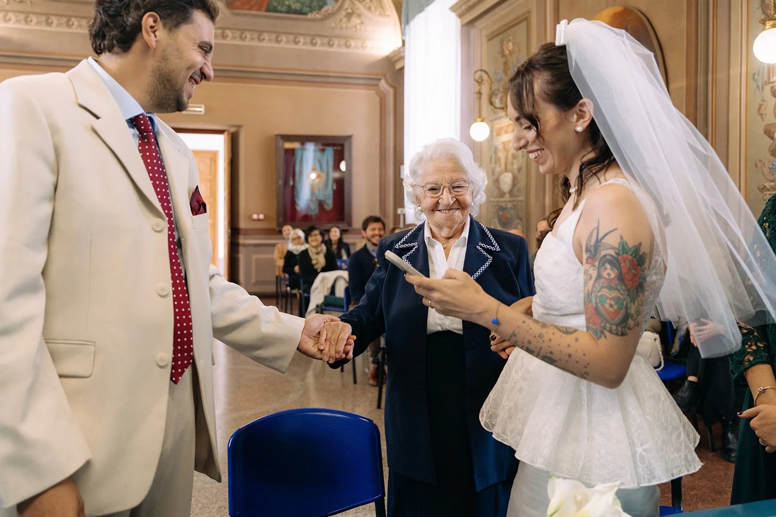 A bride in a white wedding dress and veil shaking hands with a groom in a cream suit, with an elderly woman smiling in between them during a wedding ceremony in a decorated hall, with guests in the background.