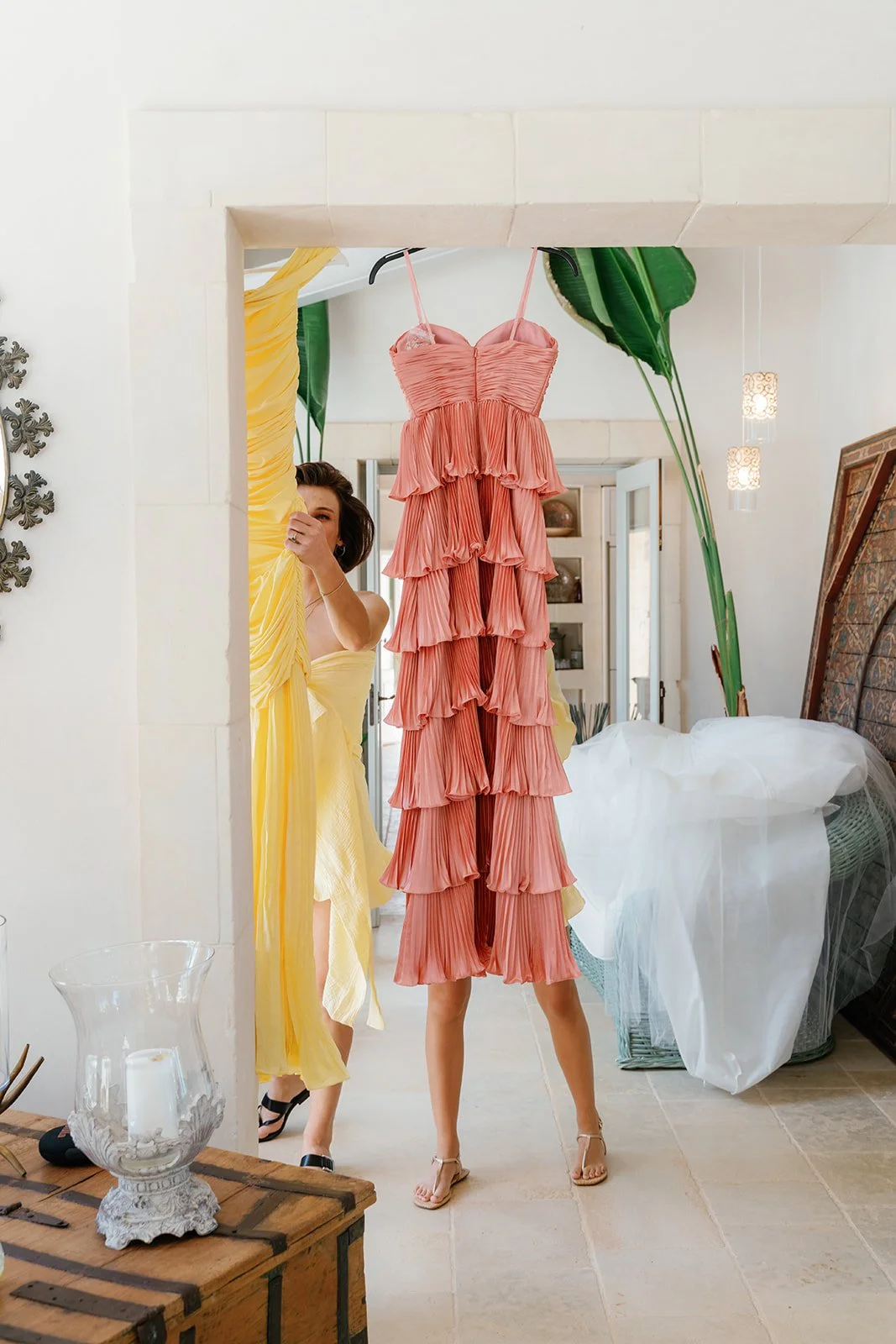 Women in yellow dresses looking at a pink pleated dress hanging in a bright, modern room with large green plants andDecor items.