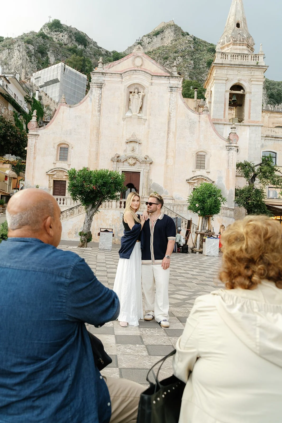 A couple is posing for a photo in front of a historic church while two other people sit nearby. The woman has blonde hair and is wearing a dark top and a long white skirt. The man has glasses and is wearing a navy blue jacket with white pants. The ch