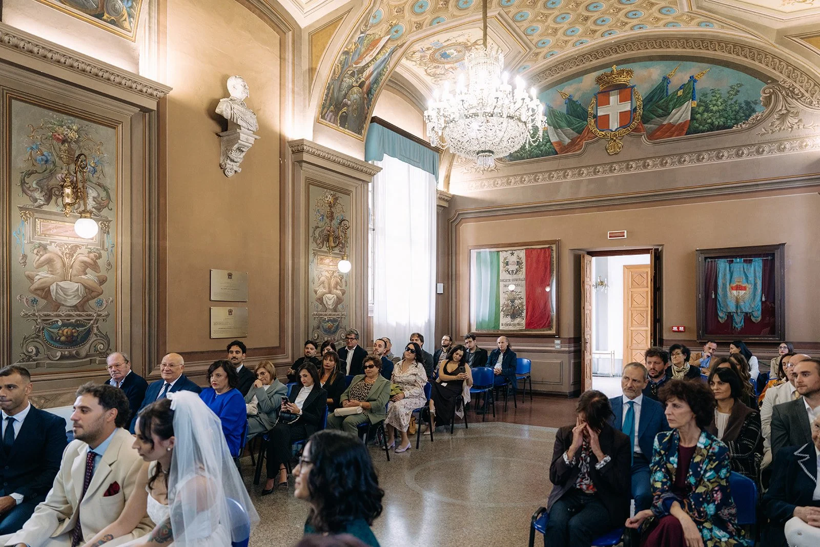 A wedding ceremony in an ornate, historical hall with high, decorated ceilings, large murals, chandeliers, and flags on the walls. The bride and groom are seated at front with guests seated behind them.