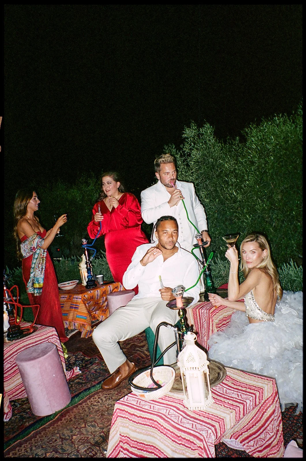 People enjoying a hookah party outdoors at night, with women in elegant dresses, a man sitting in front, and a woman in a fluffy white dress holding a martini glass.