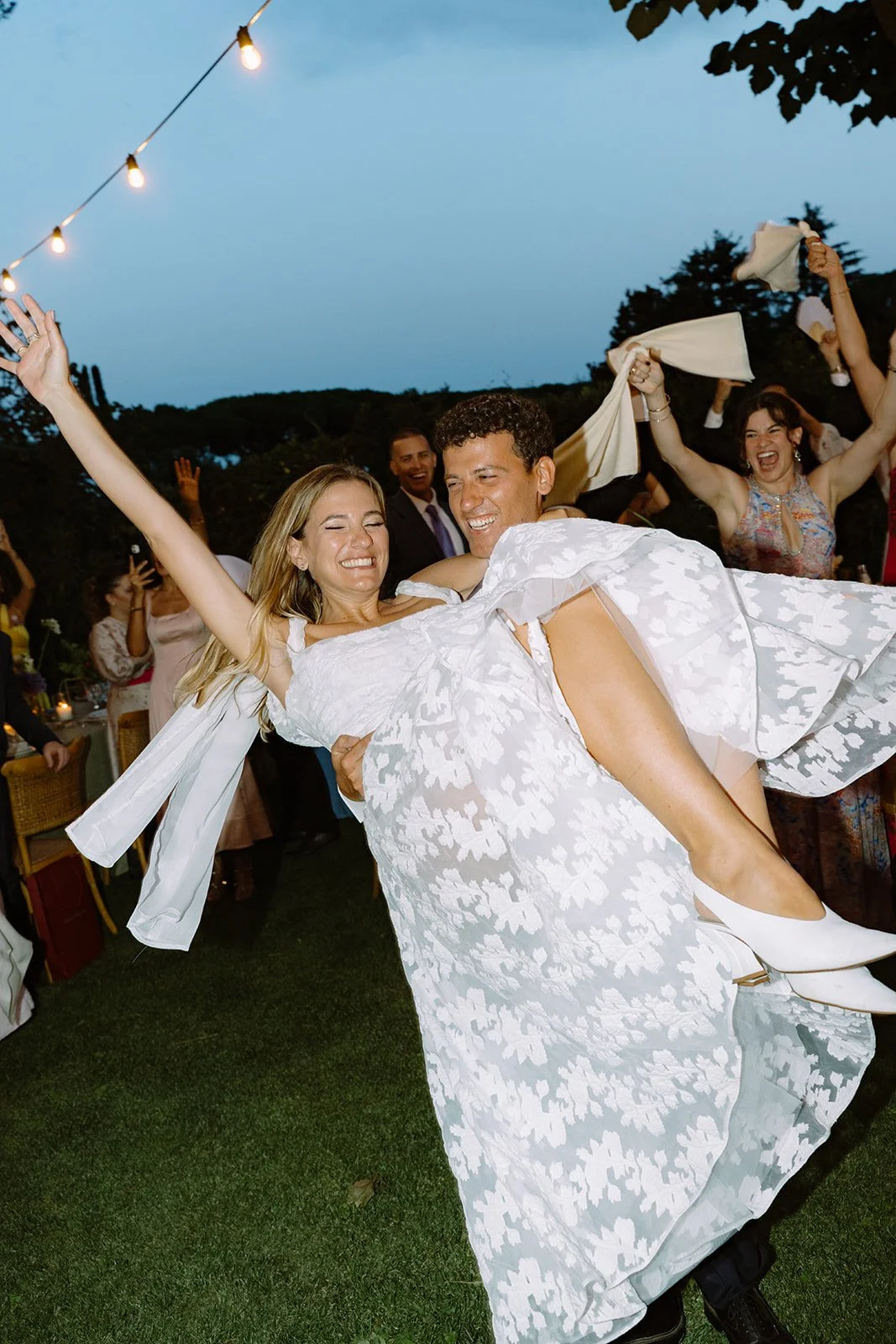 A man in a suit carrying a woman in a white dress at a celebration or wedding, surrounded by smiling people dancing and cheering outdoors at dusk.