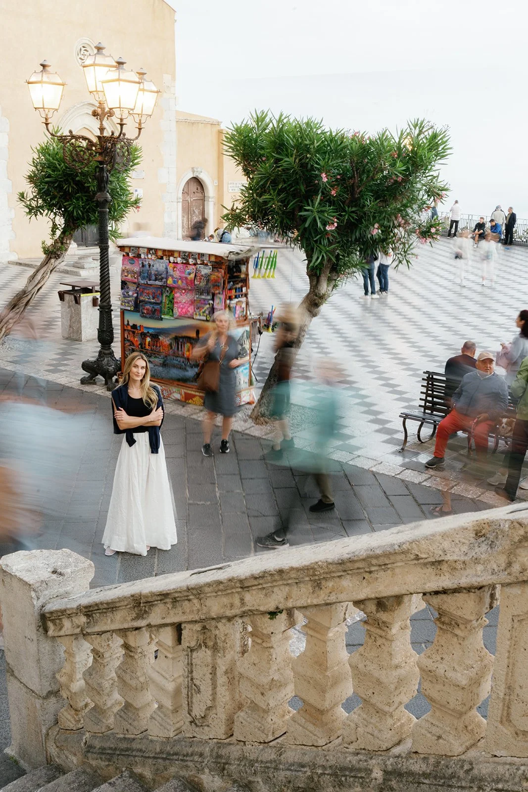 A woman with long blonde hair wearing a white skirt and black top standing on a stone staircase, surrounded by blurred people walking in a public square with a decorative streetlamp, trees, and a vendor stall in the background.
