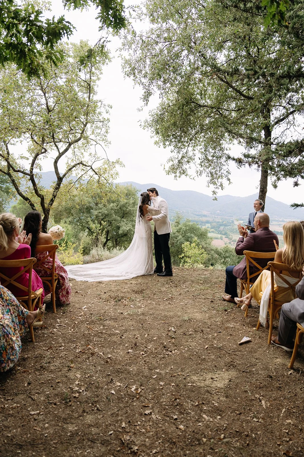 A wedding ceremony taking place outdoors on a dirt clearing surrounded by trees, with mountains in the background. The bride and groom are kissing under a large tree, with guests seated on either side.