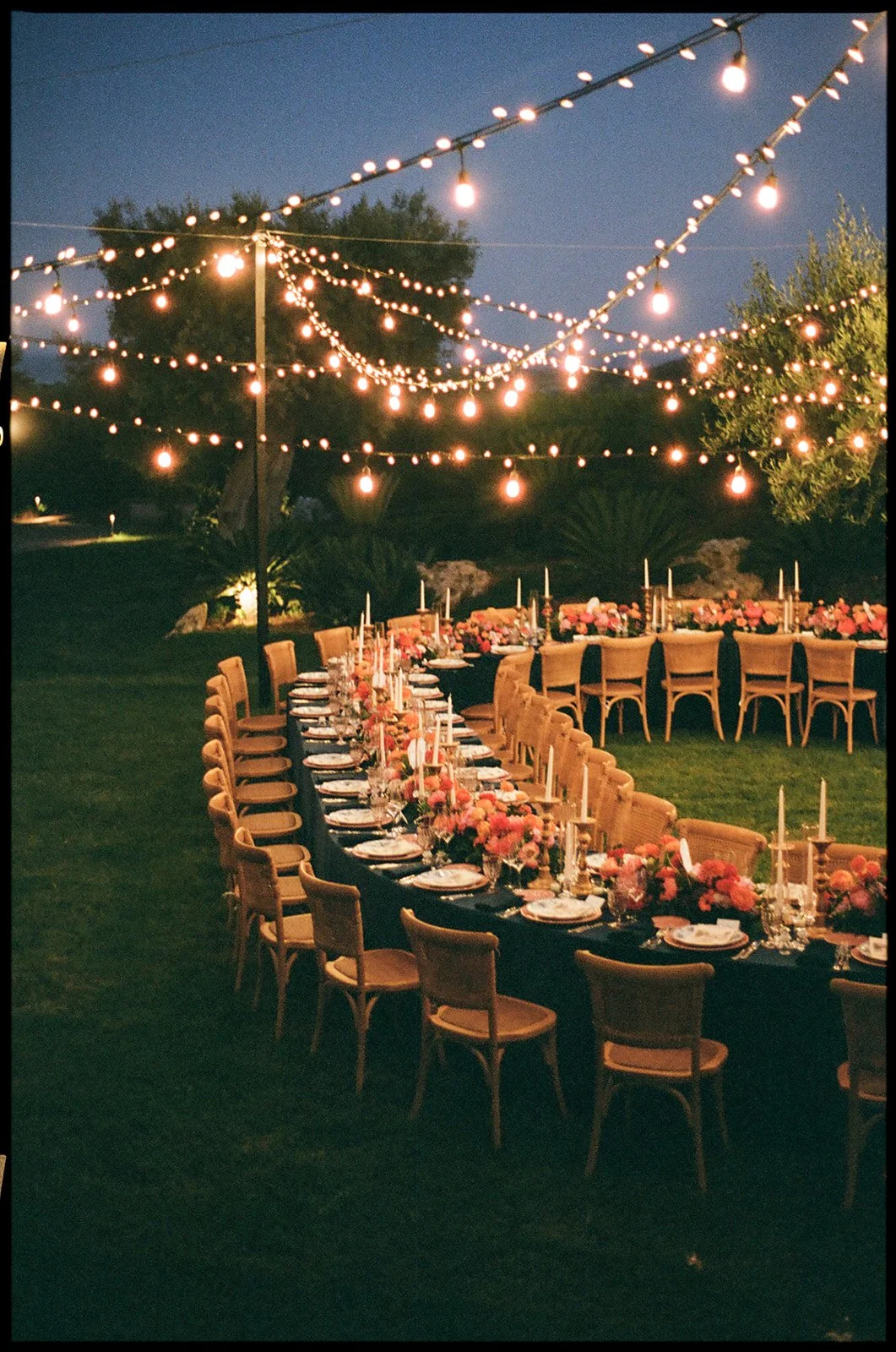 Outdoor evening dinner setup with long tables, floral centerpieces, candles, and string lights overhead in a garden setting.