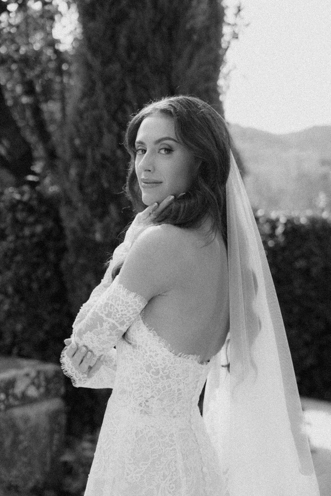 Black and white photograph of a young woman in a lacy off-the-shoulder wedding dress with a veil, standing outdoors near a large tree.