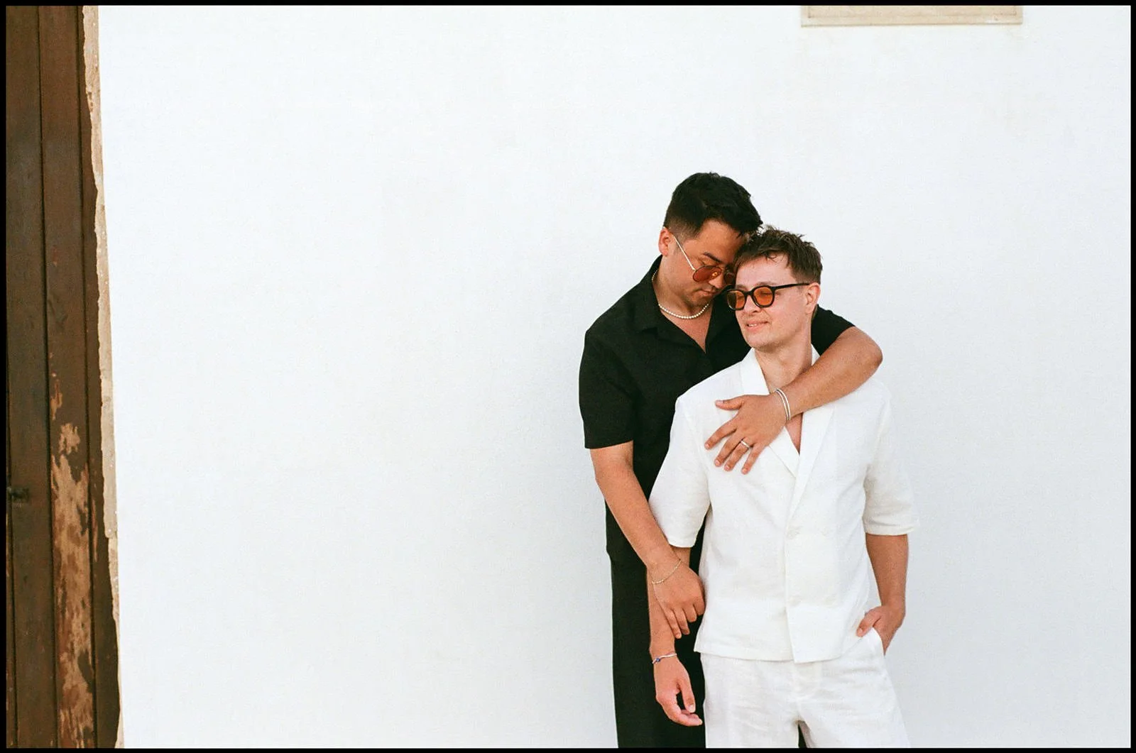 Two men standing together against a plain white wall. One man is dressed in black, wearing sunglasses and a pearl necklace, with his arm around the other man. The other man is dressed in white, also wearing sunglasses, with his hand in his pocket, an