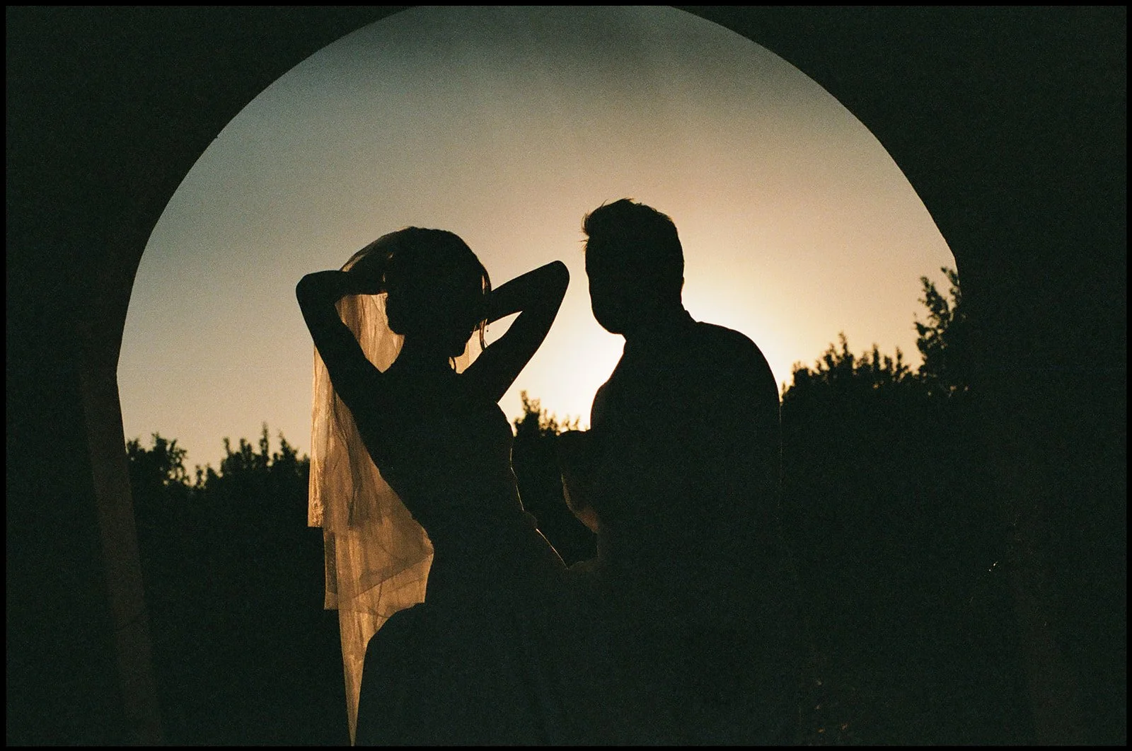 Silhouettes of a man and woman near an archway at sunset or dusk, with trees in the background. The woman is running her hands through her hair, and the man is standing close to her.
