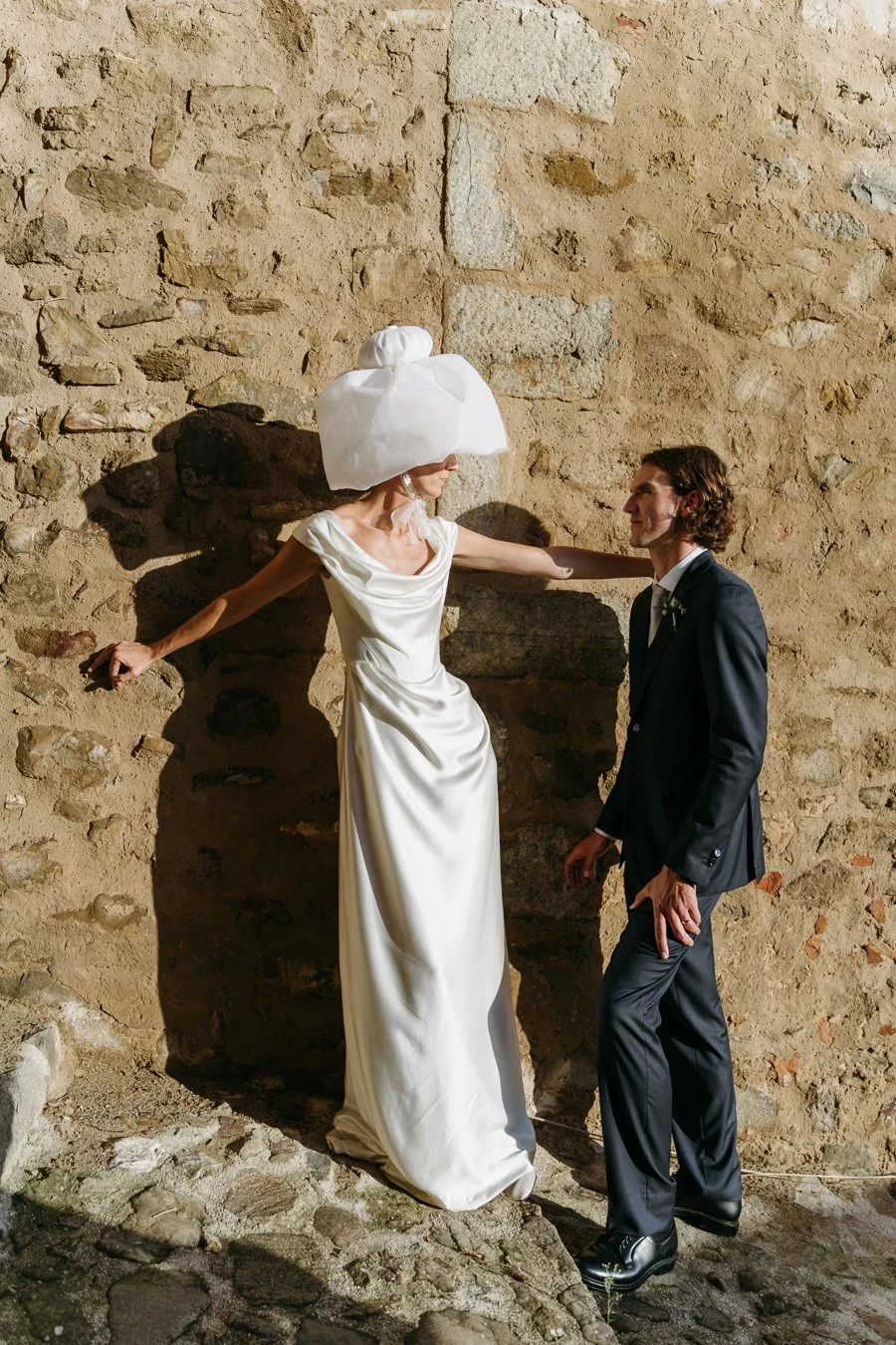 A woman in a white wedding dress with a large tulle headpiece stands against a rustic stone wall, touching her partner in a dark suit, holding a small object in her right hand.